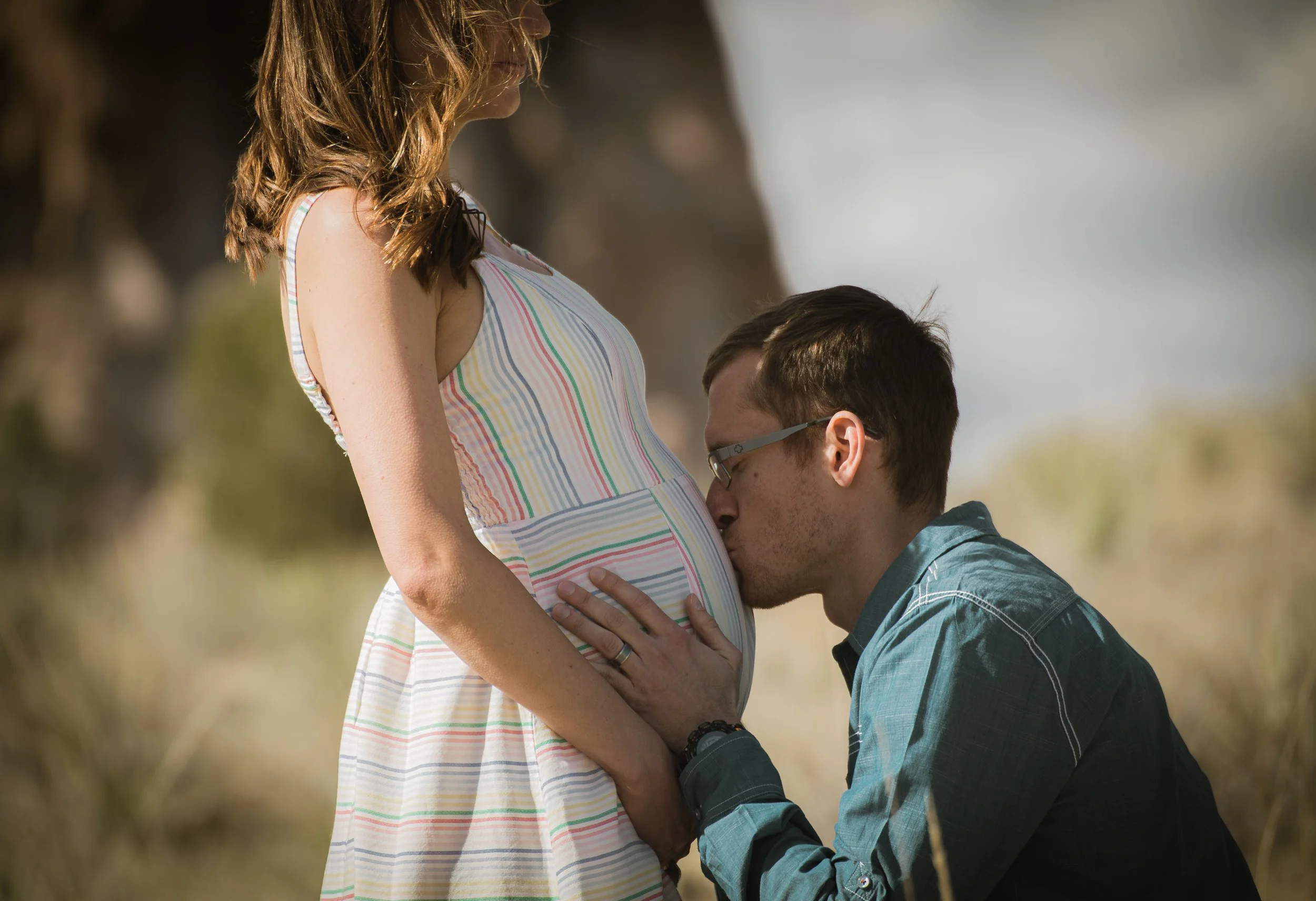 A man kisses a pregnant woman's belly outdoors in a natural setting.
