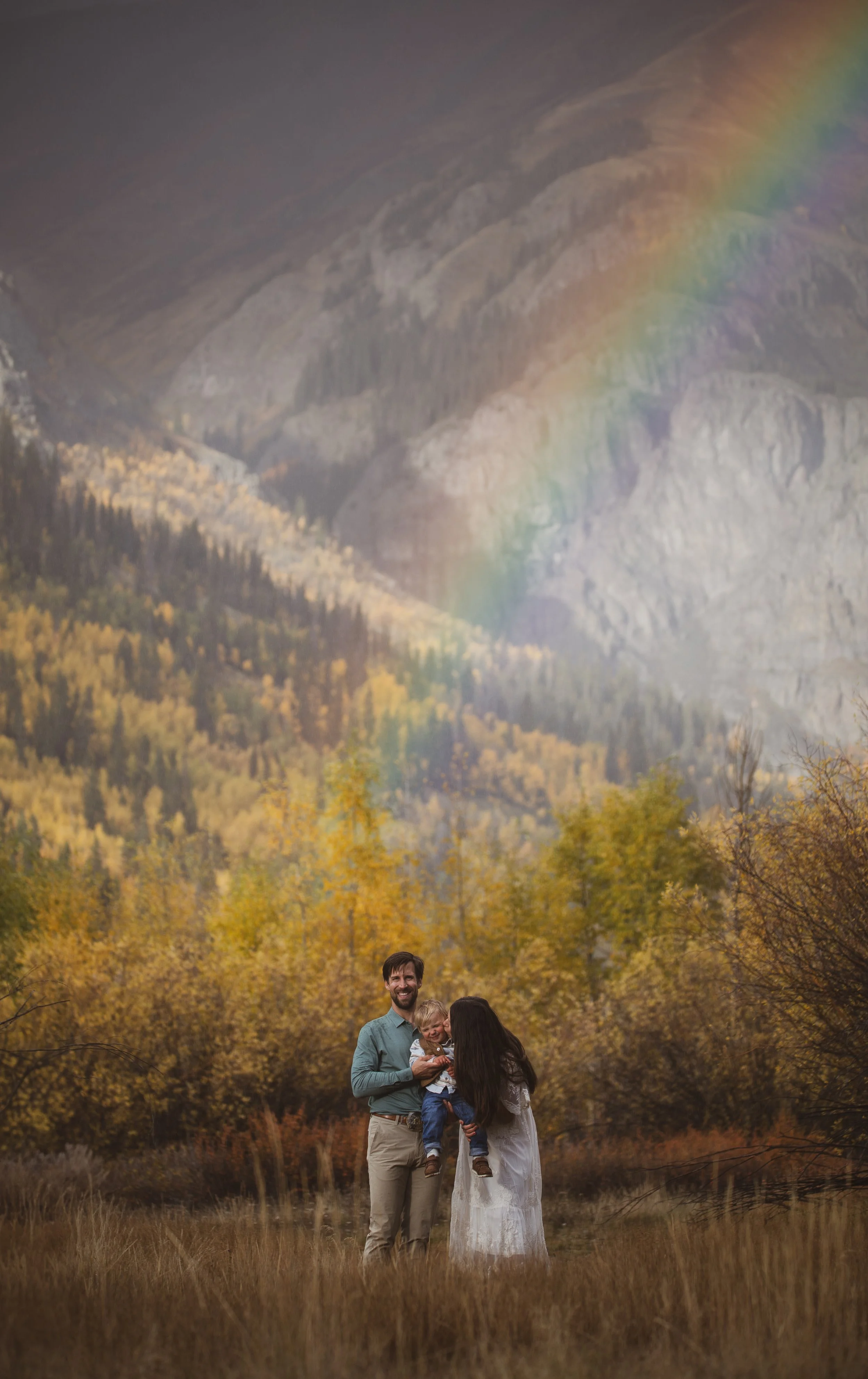 Family of three in a natural landscape with a mountain backdrop, colorful autumn trees, and a rainbow.
