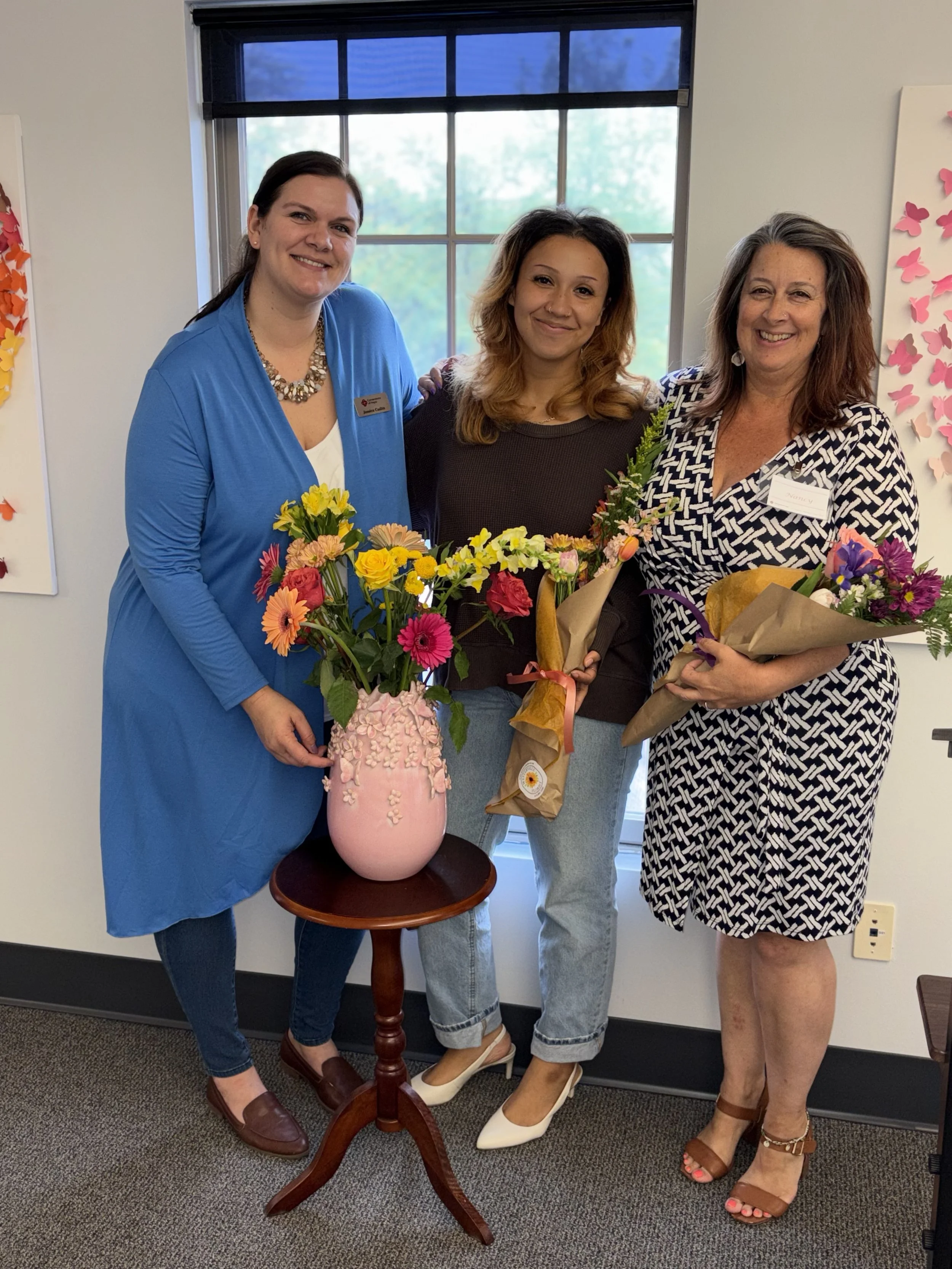 Three women standing indoors with flowers, smiling, in front of a large window and colorful butterfly artwork on the wall.