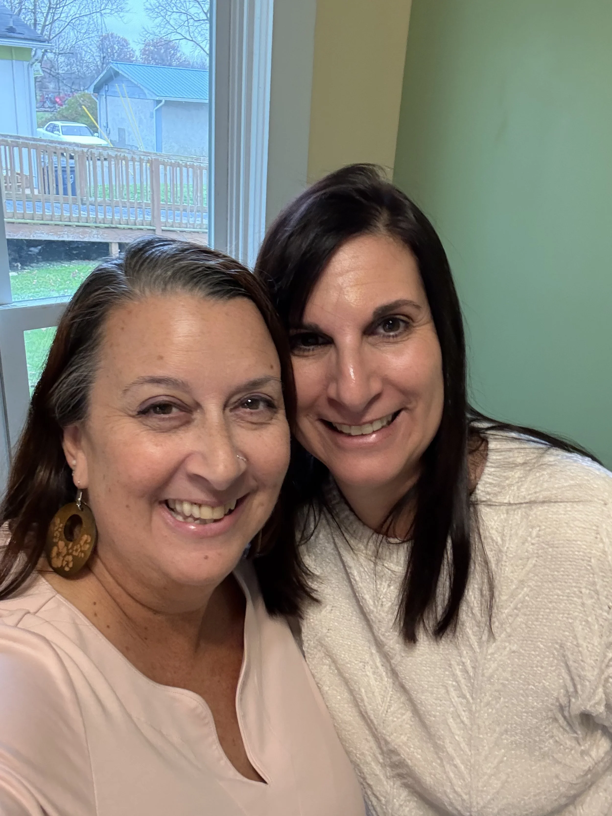 Two women smiling for a selfie in front of a window, with trees and houses visible outdoors.
