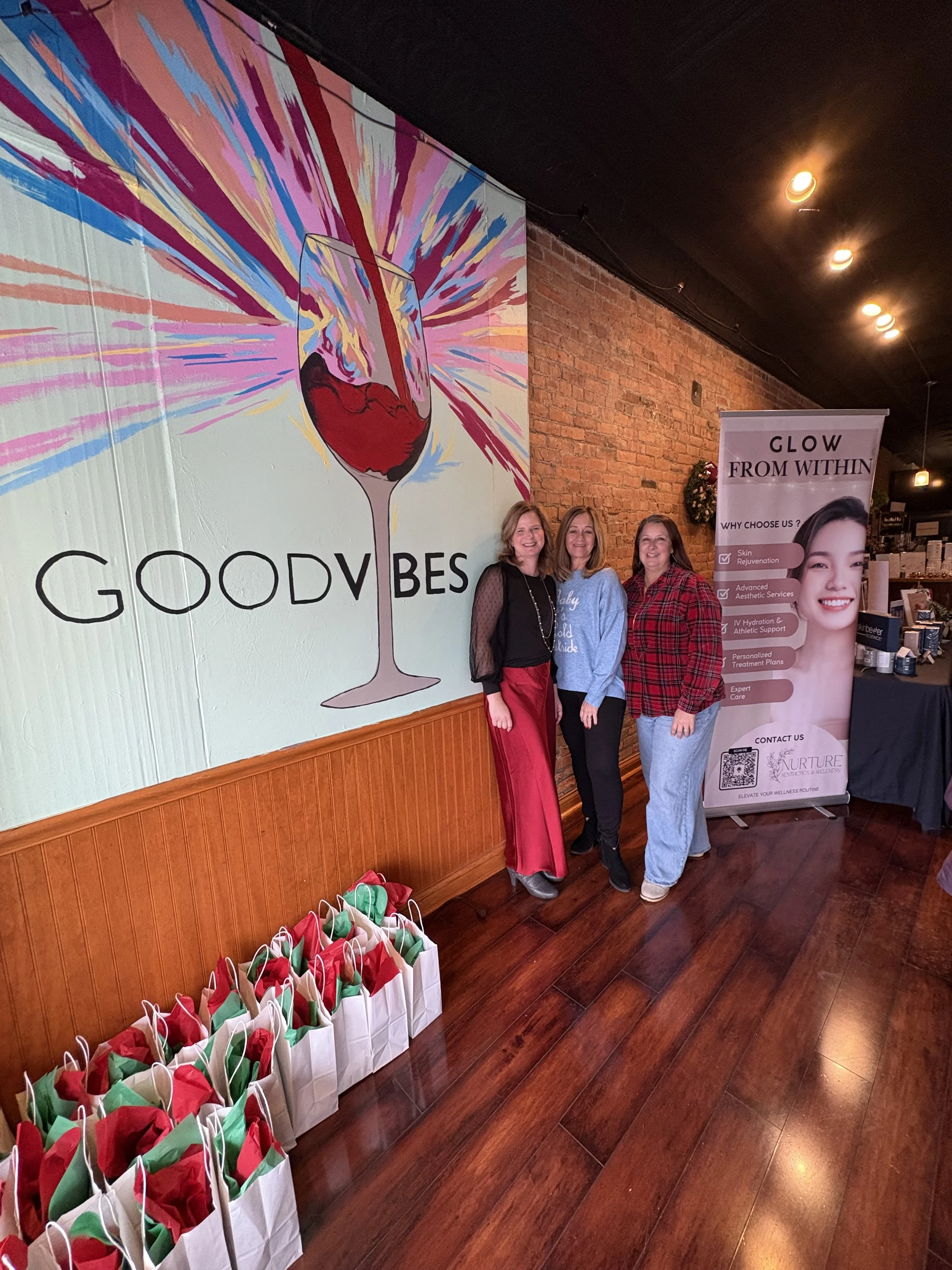 Three women standing inside a cafe or shop with a brick wall. Behind them is a large wall art of a wine glass with colorful brush strokes and the words "GOOD VIBES." To their right, a banner advertising wellness services, and in front, small gift bags are lined up on the floor.