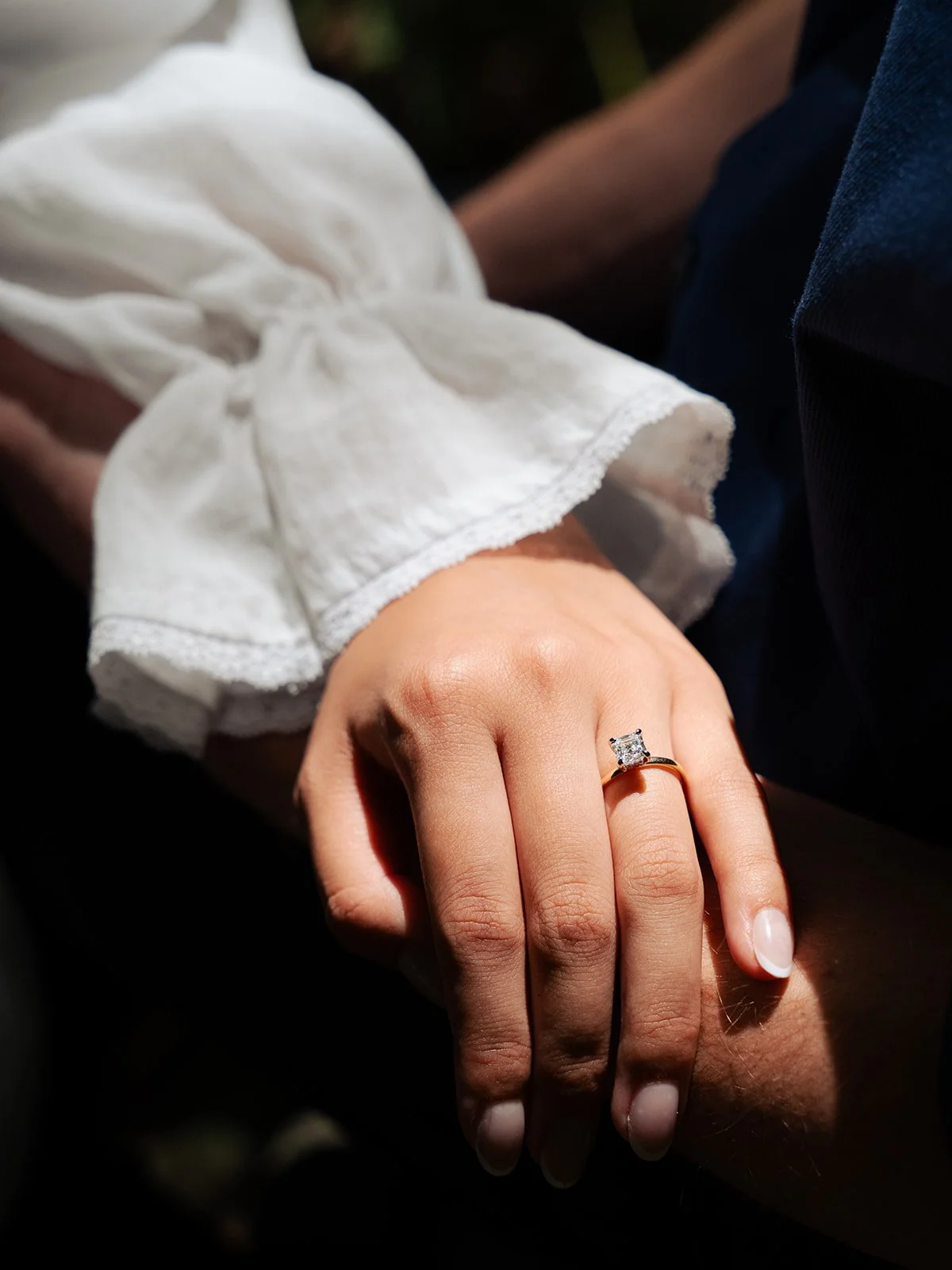 Close-up of a woman's hand resting on a person's arm, with a large diamond engagement ring on her finger, dressed in a white long-sleeve blouse.