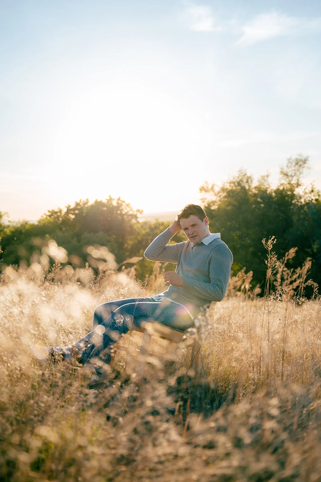 A young man sitting on a chair in a field of tall, golden grass during sunset, touching his head with one hand and looking at the camera.