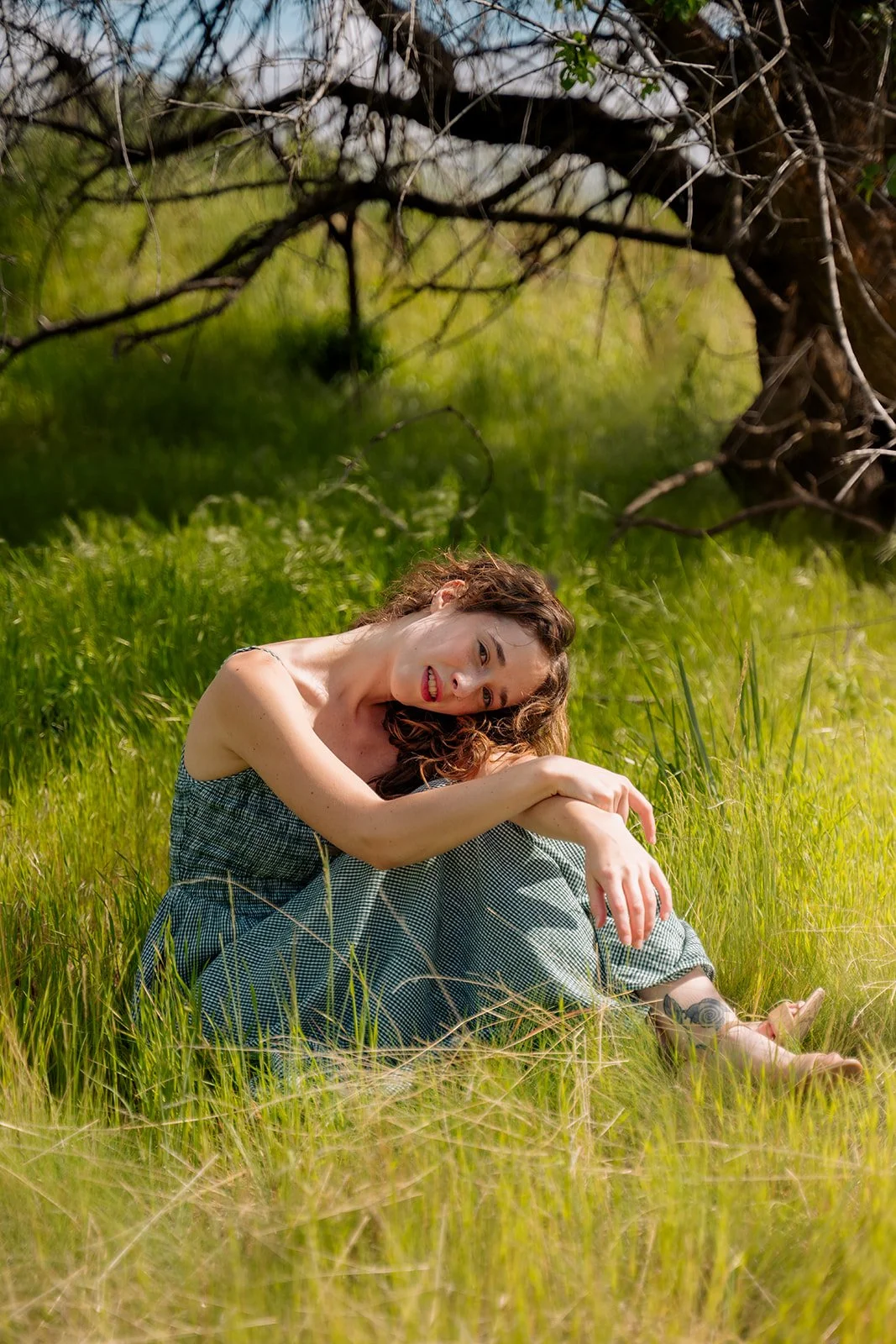 A woman with curly hair sitting on grass in a meadow with green grass and a tree in the background, smiling and leaning forward with her head tilted.