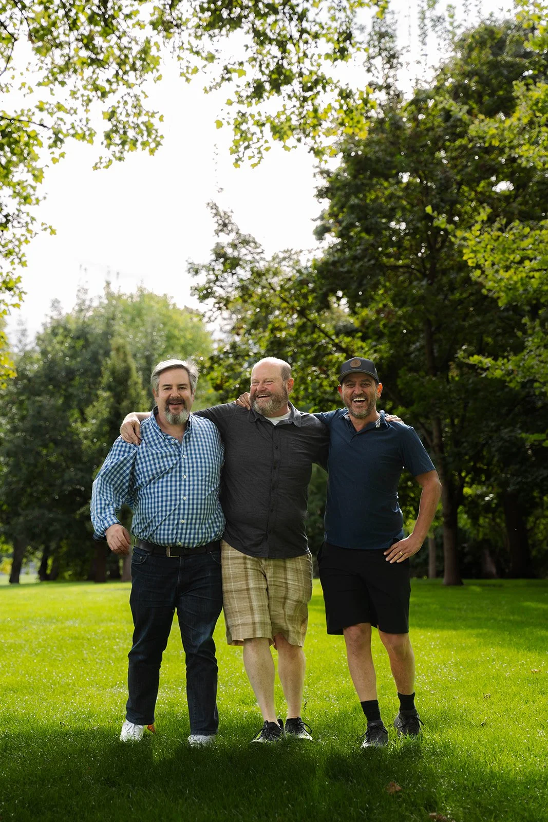 Three men standing together outdoors in a park, smiling and enjoying each other's company.