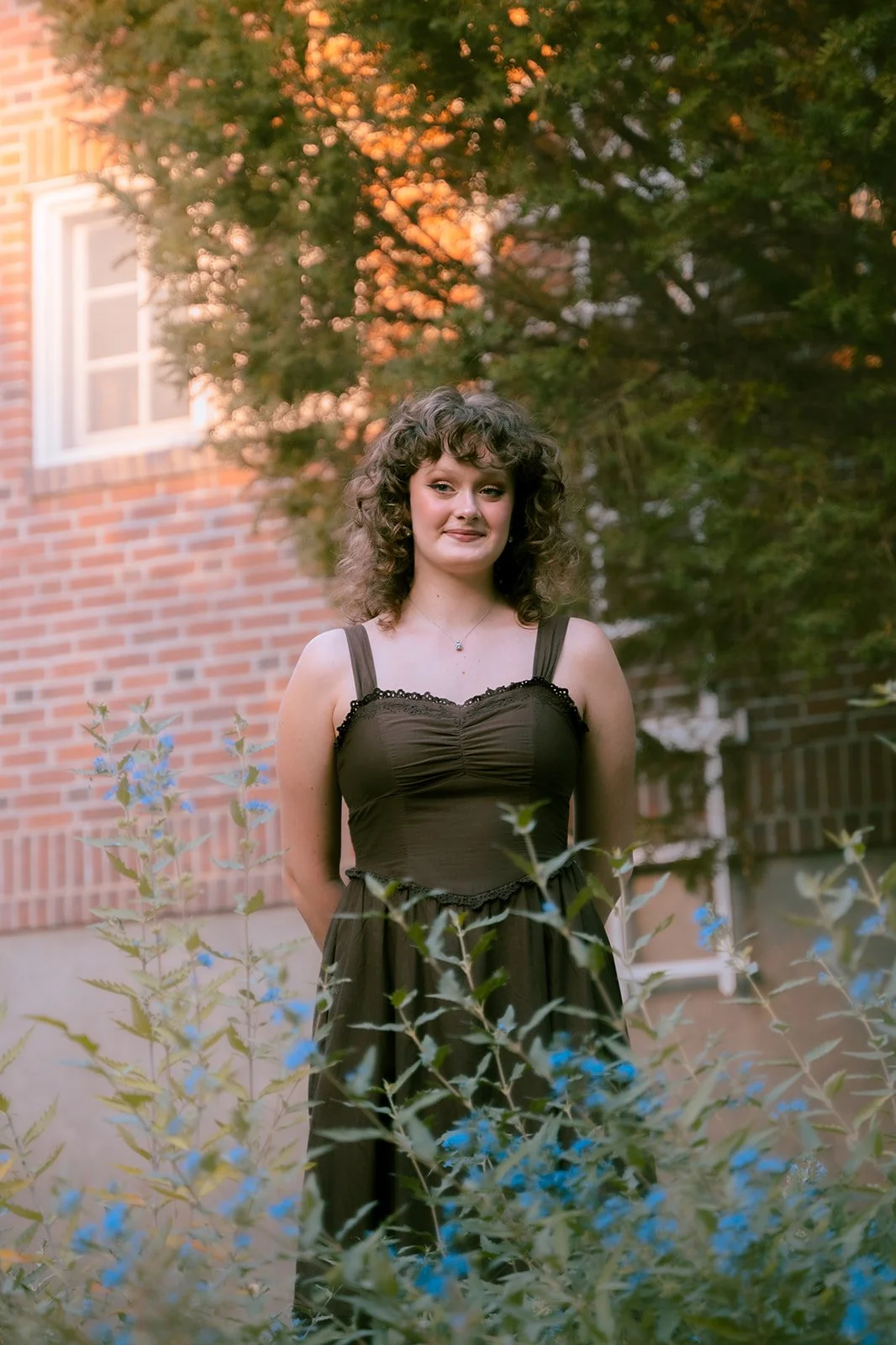 A woman with curly brown hair and a black dress standing outside behind green plants with blue flowers, in front of a brick house with white window frames, during sunset.