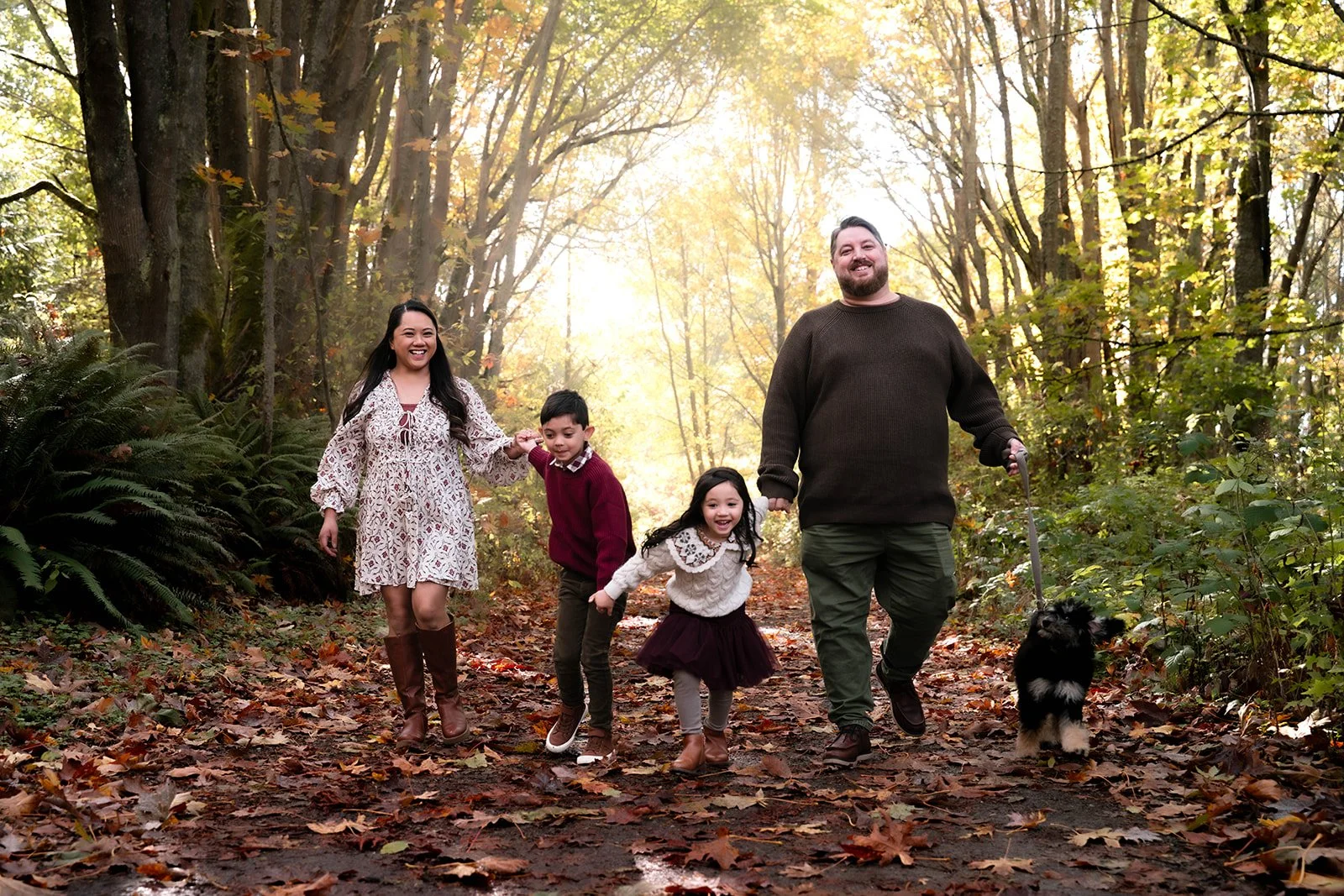Family of five walking in a sunlit autumn forest, all smiling and holding hands, with a dog on a leash, leaves scattered on the ground.