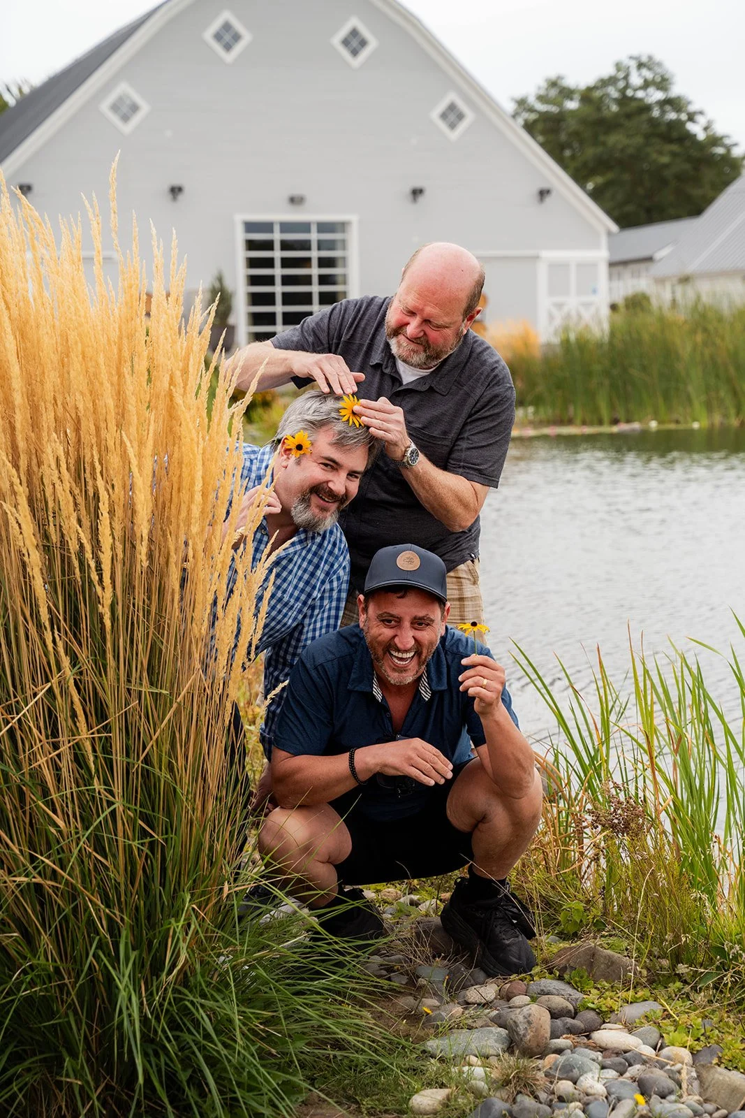 Three men by a pond, with tall yellow grass and a house in the background, are playingfully holding flowers to each other's heads and smiling.