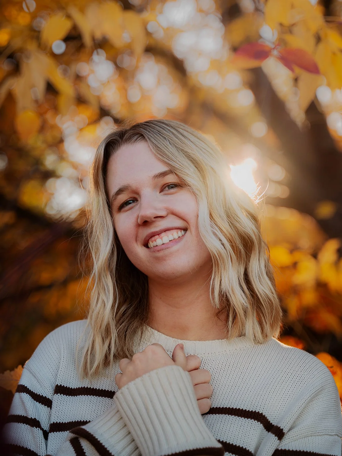 A young woman with blonde, wavy hair smiling outdoors during autumn, with sunlight filtering through yellow leaves in the background.