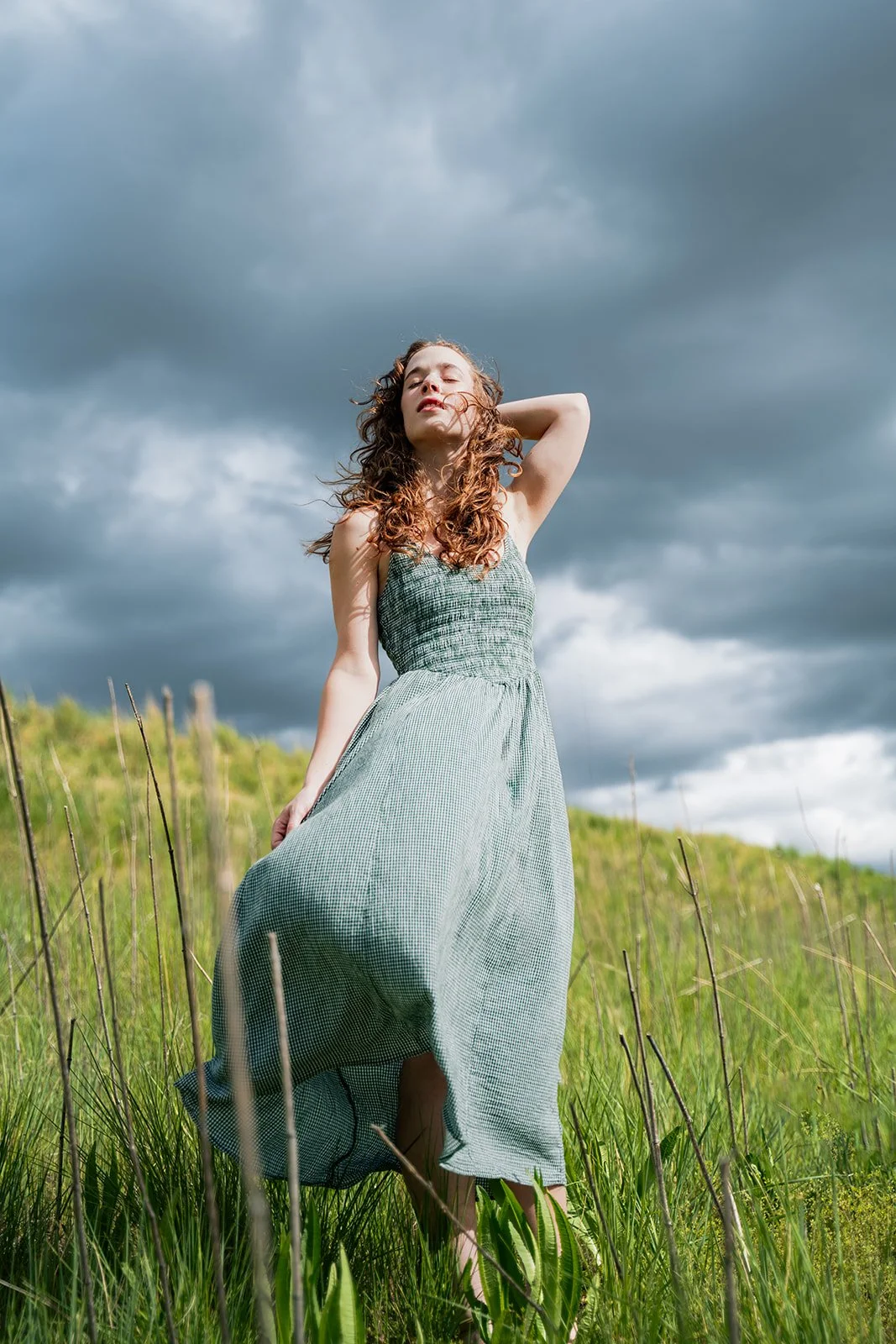 A young woman with curly red hair in a green dress standing in a grassy field under a cloudy sky, with her eyes closed and one hand behind her head.