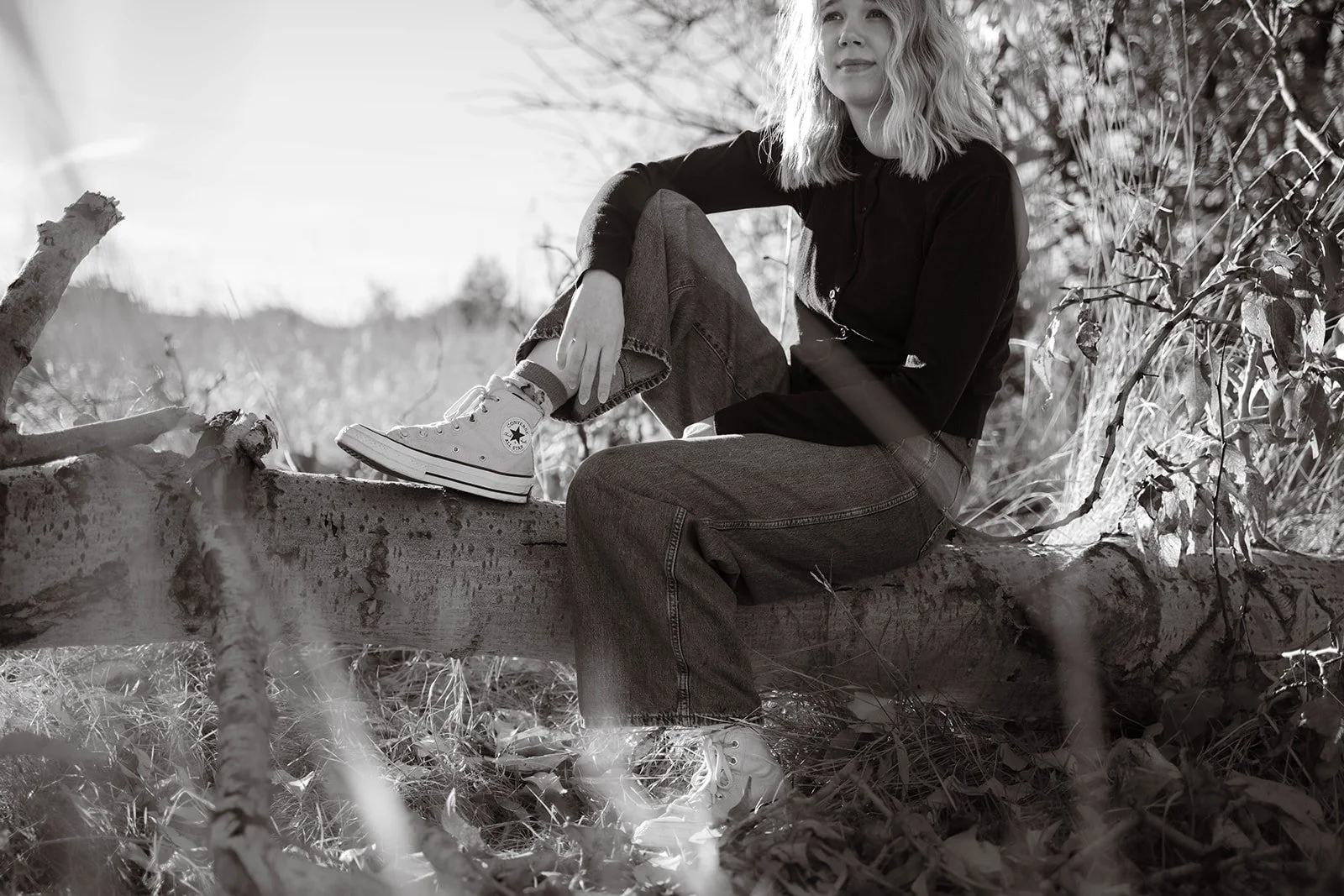 A young woman sitting on a fallen tree in a natural outdoor setting, wearing a black long-sleeve shirt, loose jeans, and white Converse sneakers, with a relaxed and contemplative expression.
