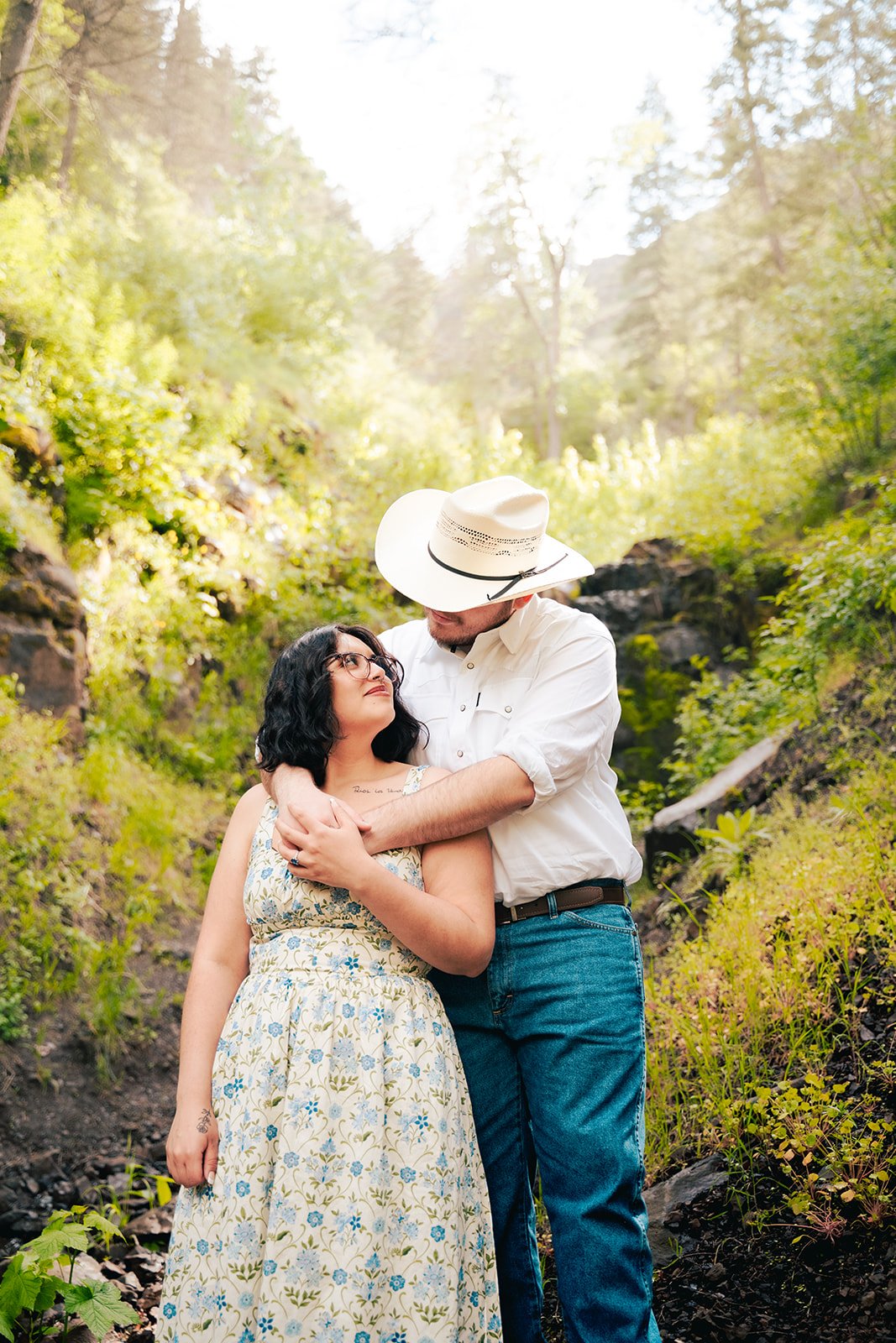 A couple embracing outdoors in a lush, green forest with sunlight streaming through trees.