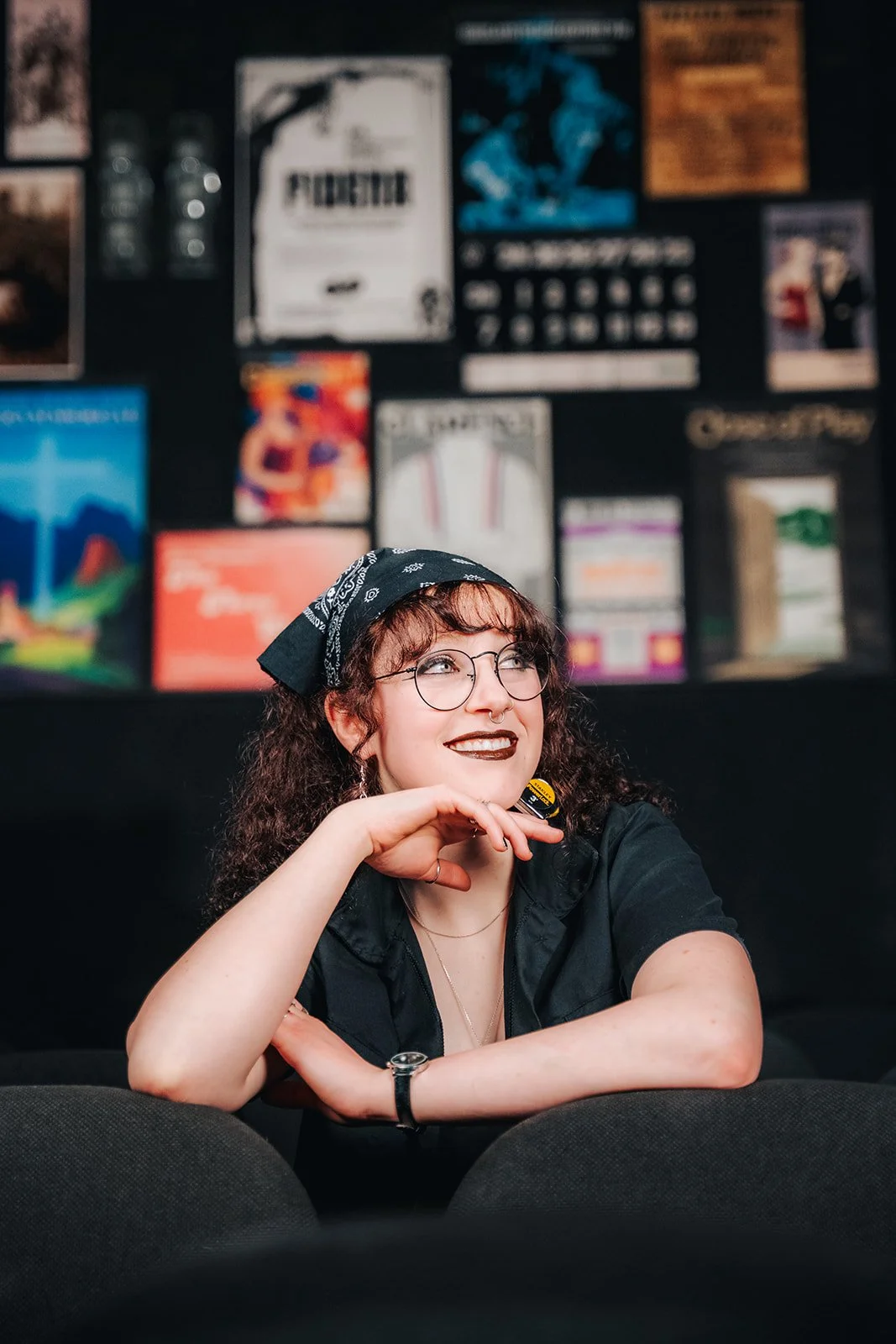 A woman with curly hair, glasses, and dark lipstick, sitting with her chin resting on her hand, smiling in a room with a wall decorated with colorful posters.