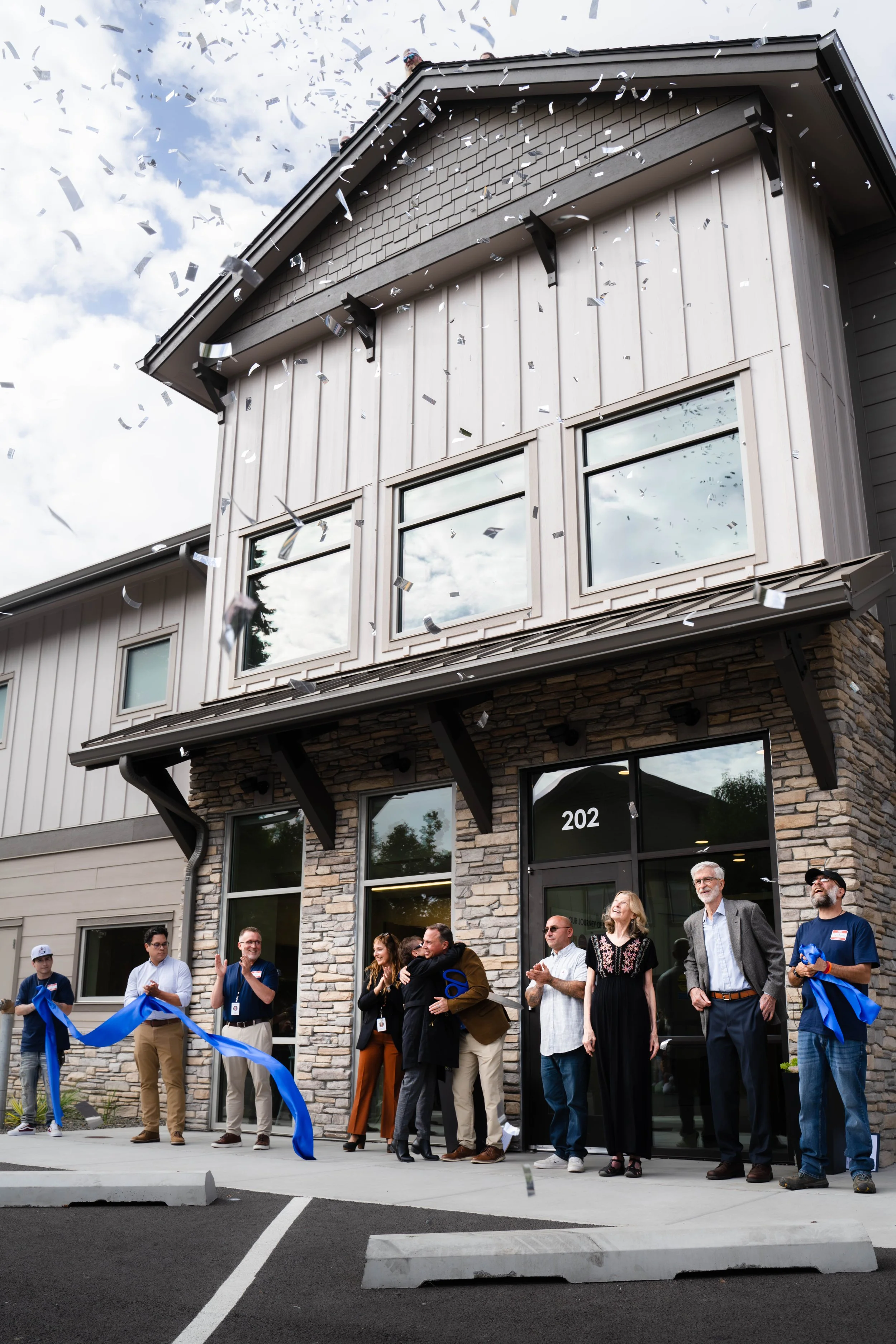 People celebrating a ribbon-cutting ceremony outside a new building with the number 202 on the door. Confetti is falling from above.