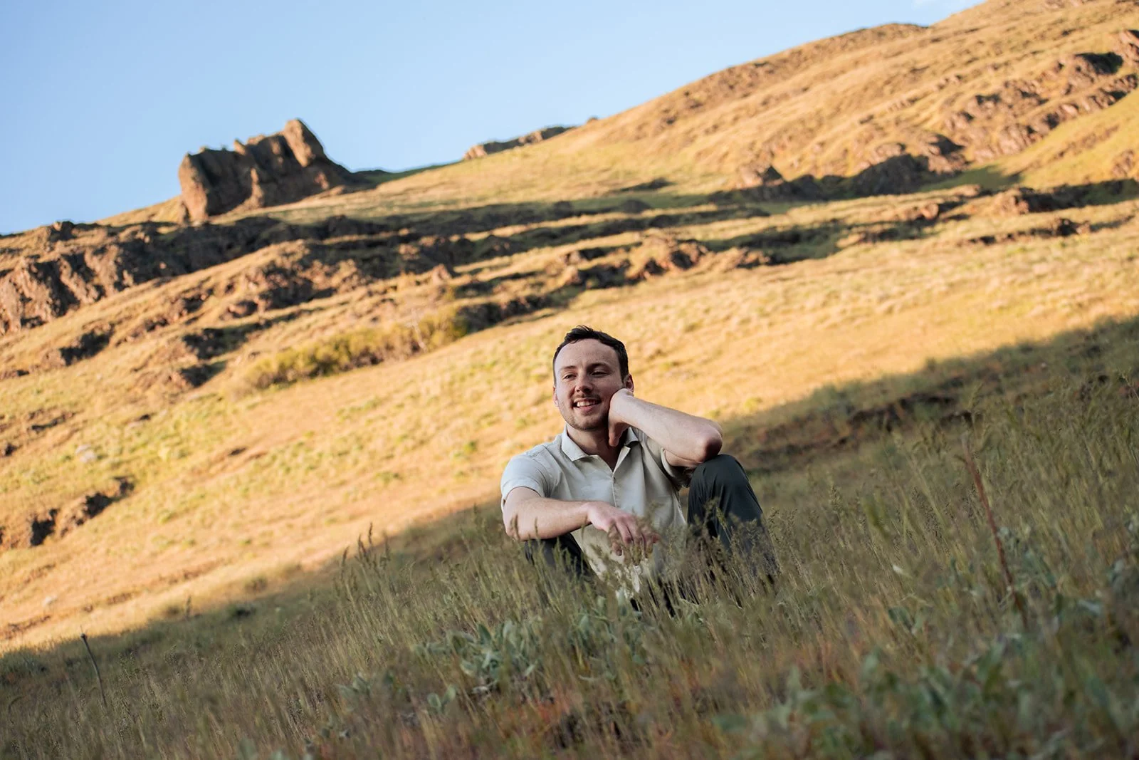 A man sitting in tall grass outdoors with a mountainous landscape in the background, smiling and resting his head on his hand.