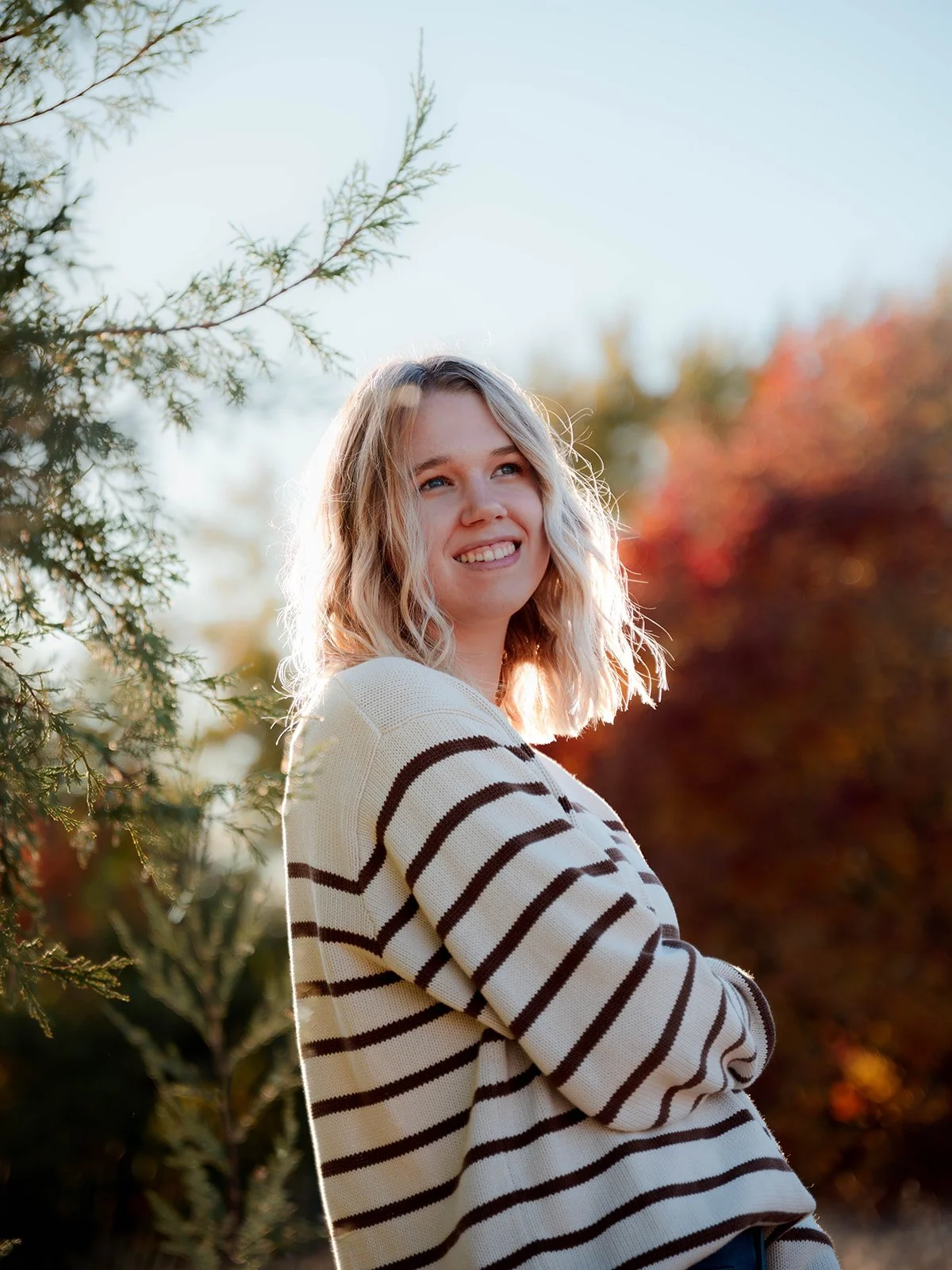 A young woman with shoulder-length blonde wavy hair smiling outdoors in autumn, wearing a white sweater with black horizontal stripes, with trees and fall foliage in the background.