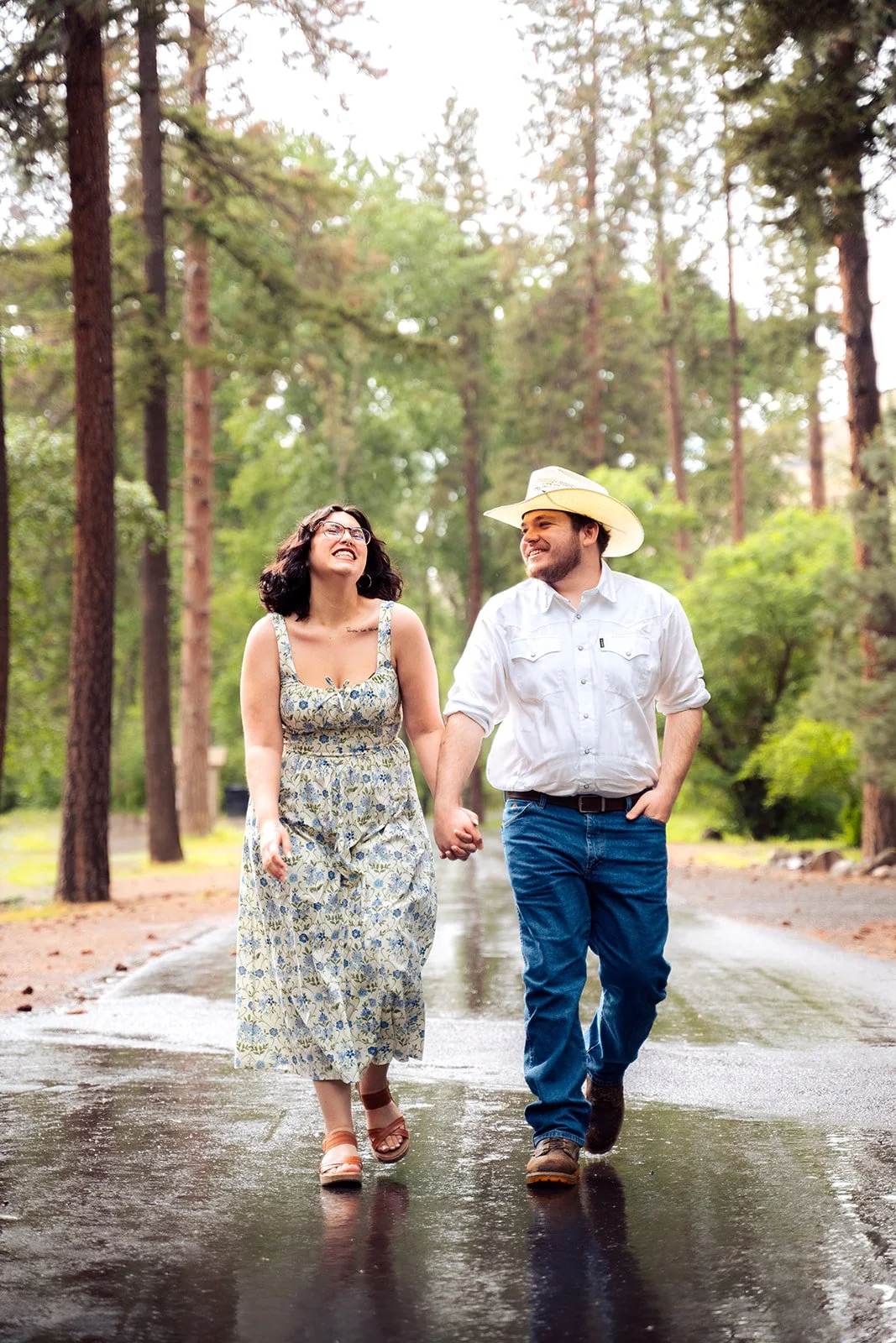 A couple walking hand in hand on a wet park trail surrounded by tall trees, smiling and enjoying each other's company.