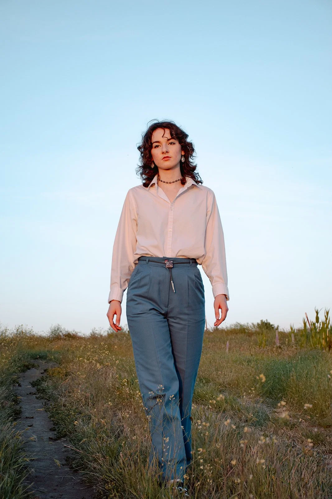 A young woman with curly hair walks along a grassy path during sunset, dressed in a white shirt and blue pants, against a clear blue sky.