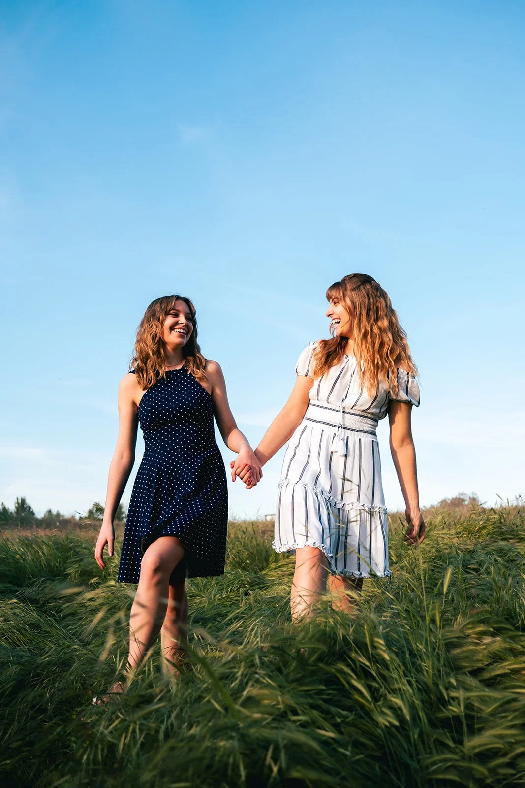 Two women holding hands and walking through a grassy field, smiling at each other under a clear blue sky.