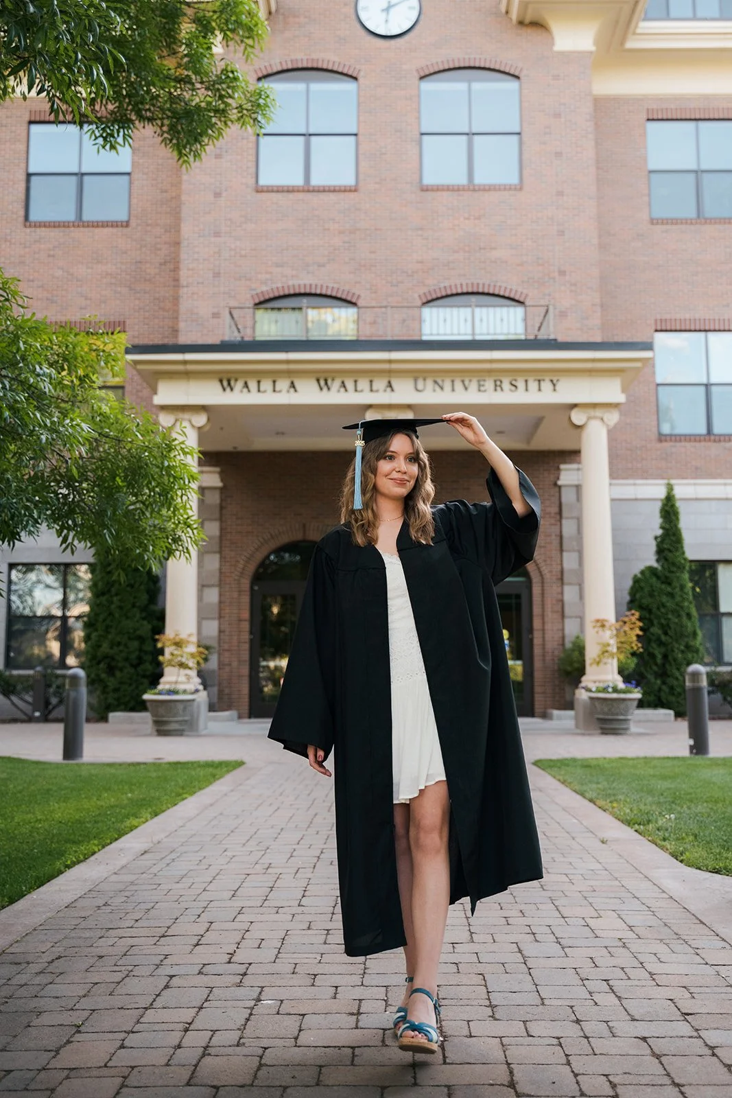 A woman in a black graduation gown and cap standing on a brick path outside Walla Walla University, adjusting her cap with one hand, with a brick university building in the background.
