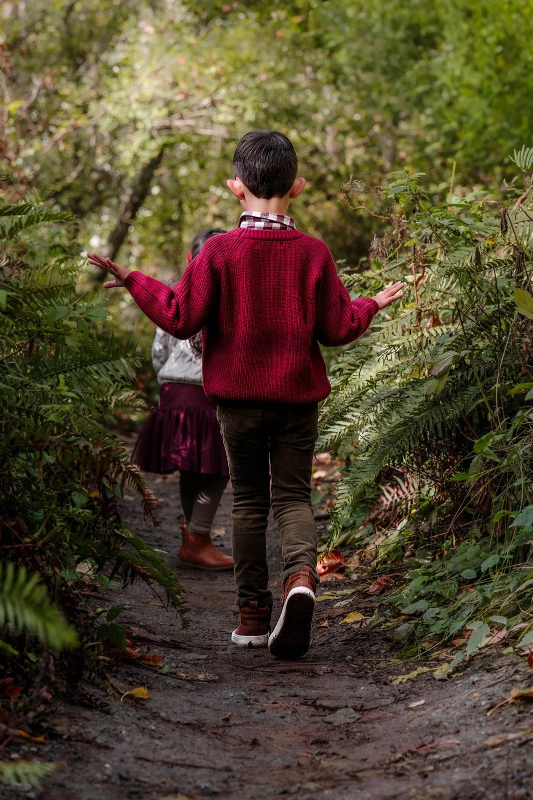 A young boy with short dark hair walking on a narrow dirt trail through a forest with green foliage, wearing a red sweater, brown pants, and brown boots, with his arms outstretched.