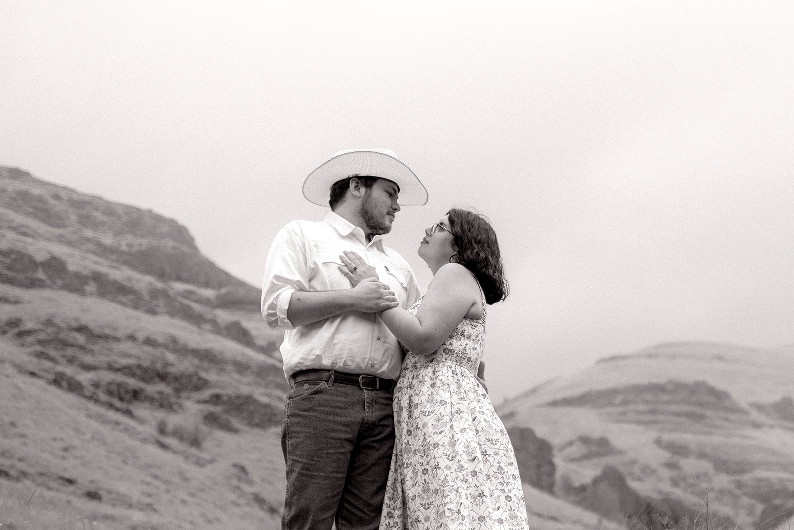 A black and white photo of a man and woman standing close together outdoors, with hills in the background. The man is wearing a cowboy hat and a button-up shirt, while the woman is in a sleeveless floral dress. They are gazing into each other's eyes,