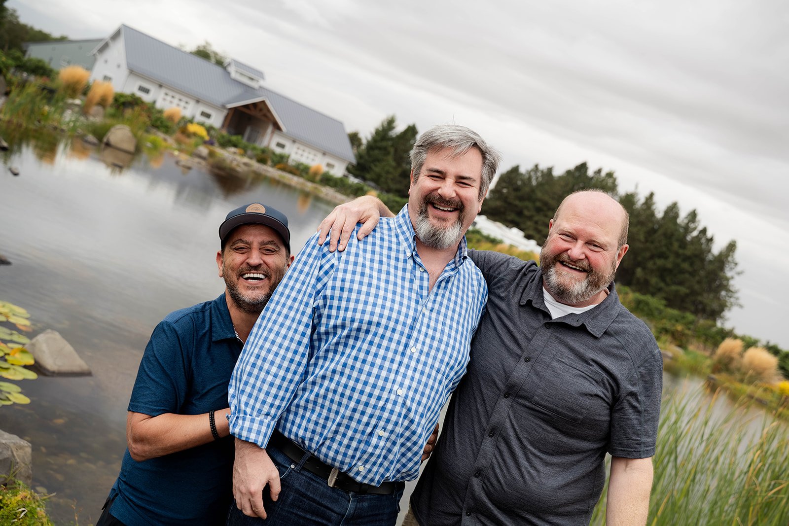 Three men smiling, standing outdoors near a body of water with a house and trees in the background.