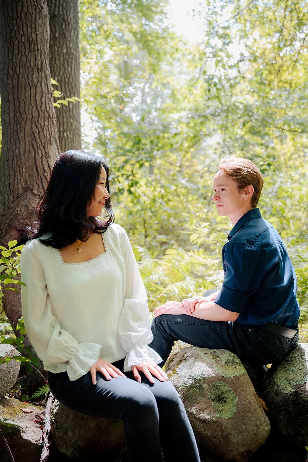 A woman and a man sitting on rocks in a forest, facing each other, with sunlight filtering through trees in the background.