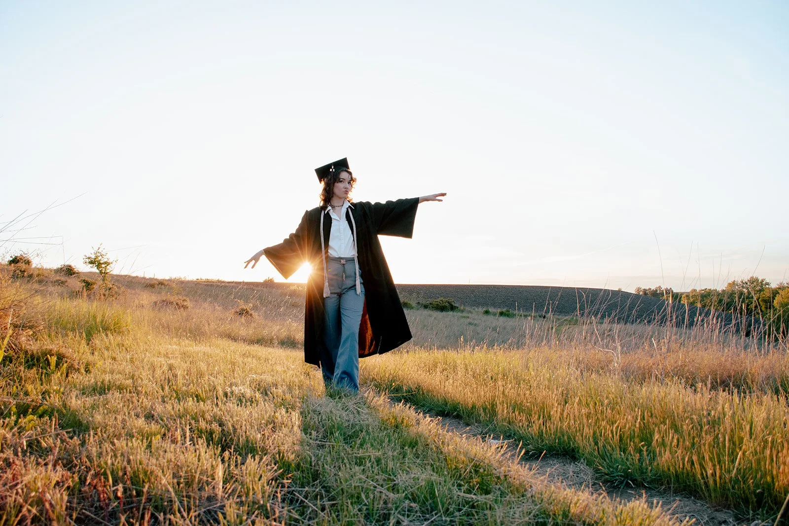 A woman in graduation cap and gown balancing on a narrow dirt path in a field with tall grass at sunset.