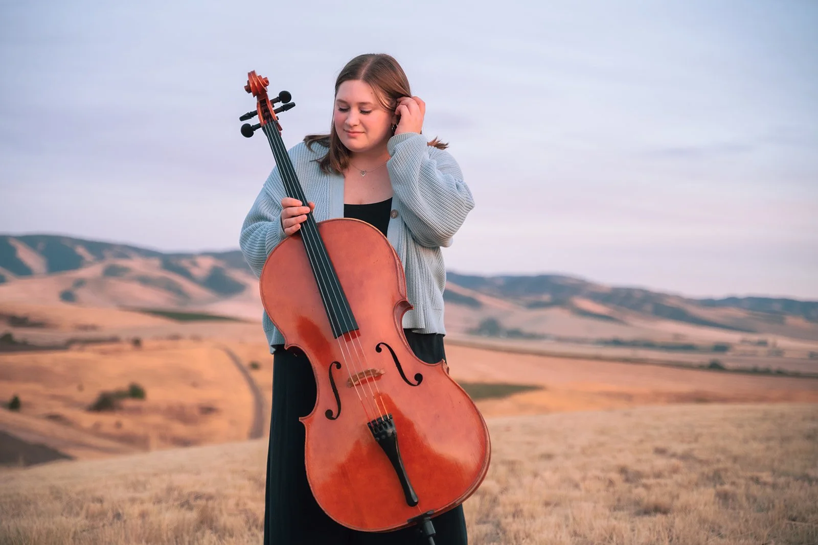 A young woman standing in a dry, hilly landscape holding a cello, with her eyes closed and a serene expression.