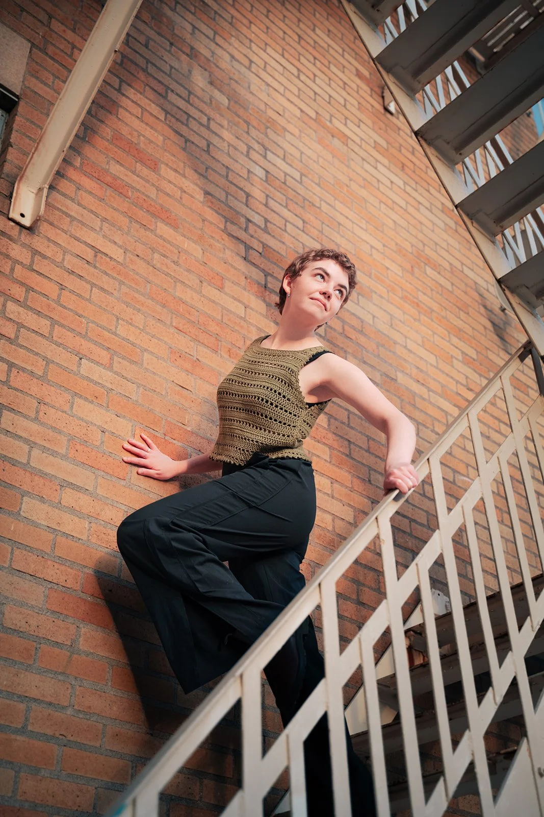 A young woman with short hair poses on a spiral staircase outdoors, leaning against a brick wall, looking upward and to the side.
