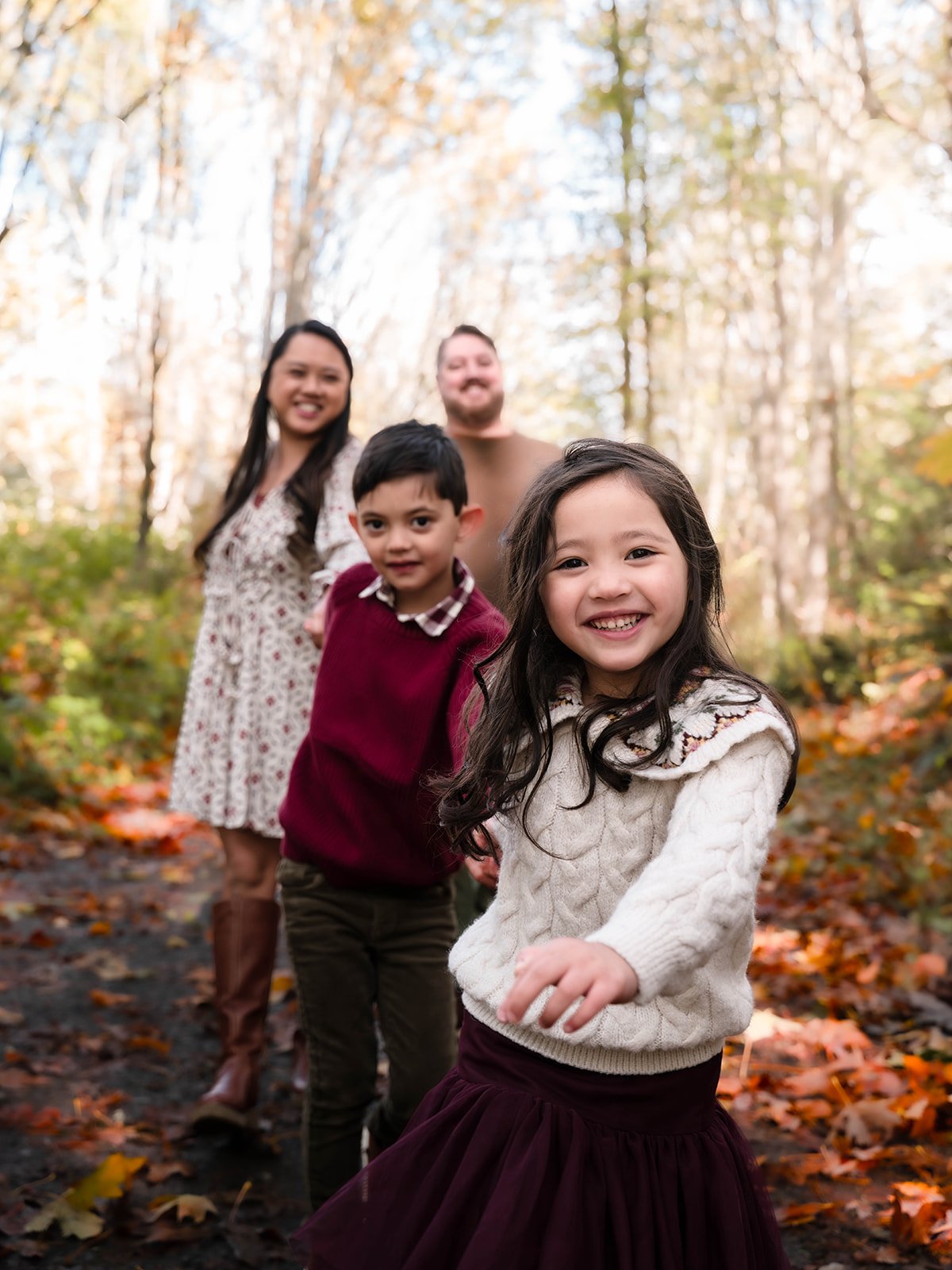 A family of five, including two young girls, a boy, and two adults, walking outdoors on a fall day through a forest with fallen leaves on the ground, smiling and enjoying the weather.