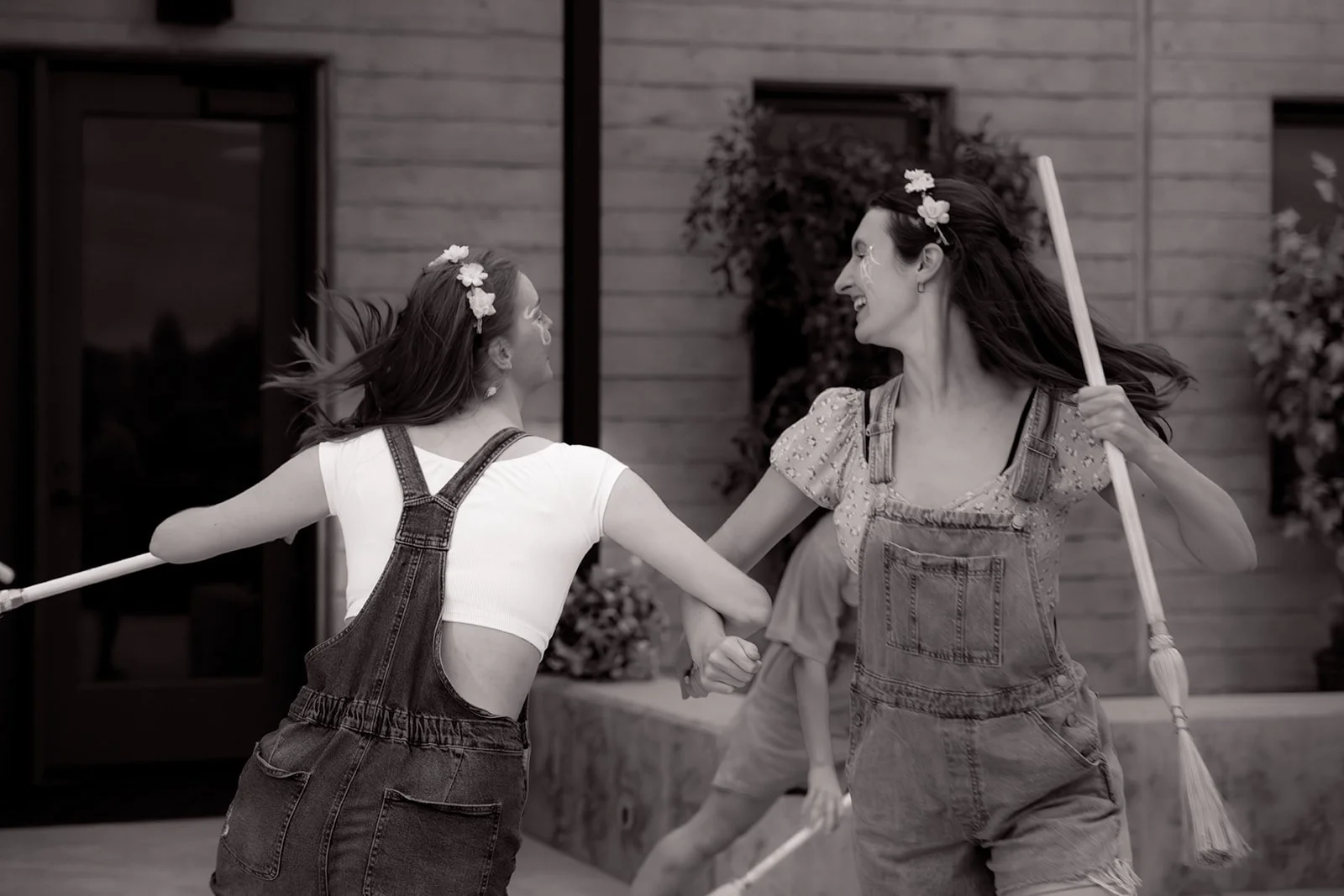 Two women wearing overalls and floral headbands, holding brooms, laughing and dancing outside a house.