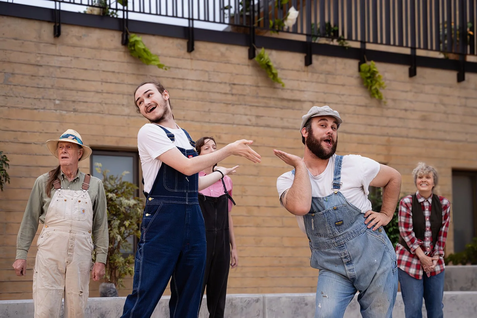 A group of five people, six including a child, engaging in a theatrical performance outdoors. They wear casual and work clothing, with some in overalls, and express various emotions and humorous poses against a modern building with plants.