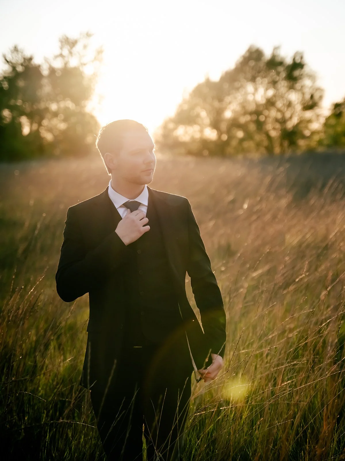 A young man in a black suit, white shirt, and black tie standing in a field at sunset, adjusting his tie, with trees in the background and sunlight creating a halo effect around his head.