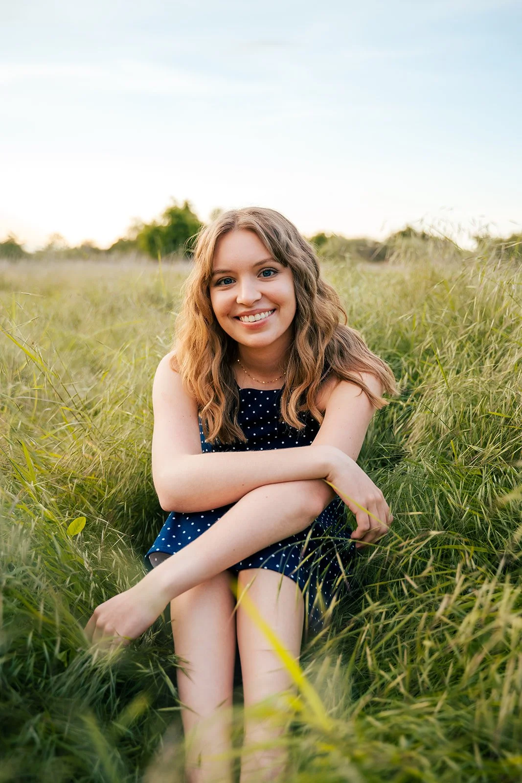 A young woman with wavy brown hair sitting in a grassy field, smiling at the camera during sunset.