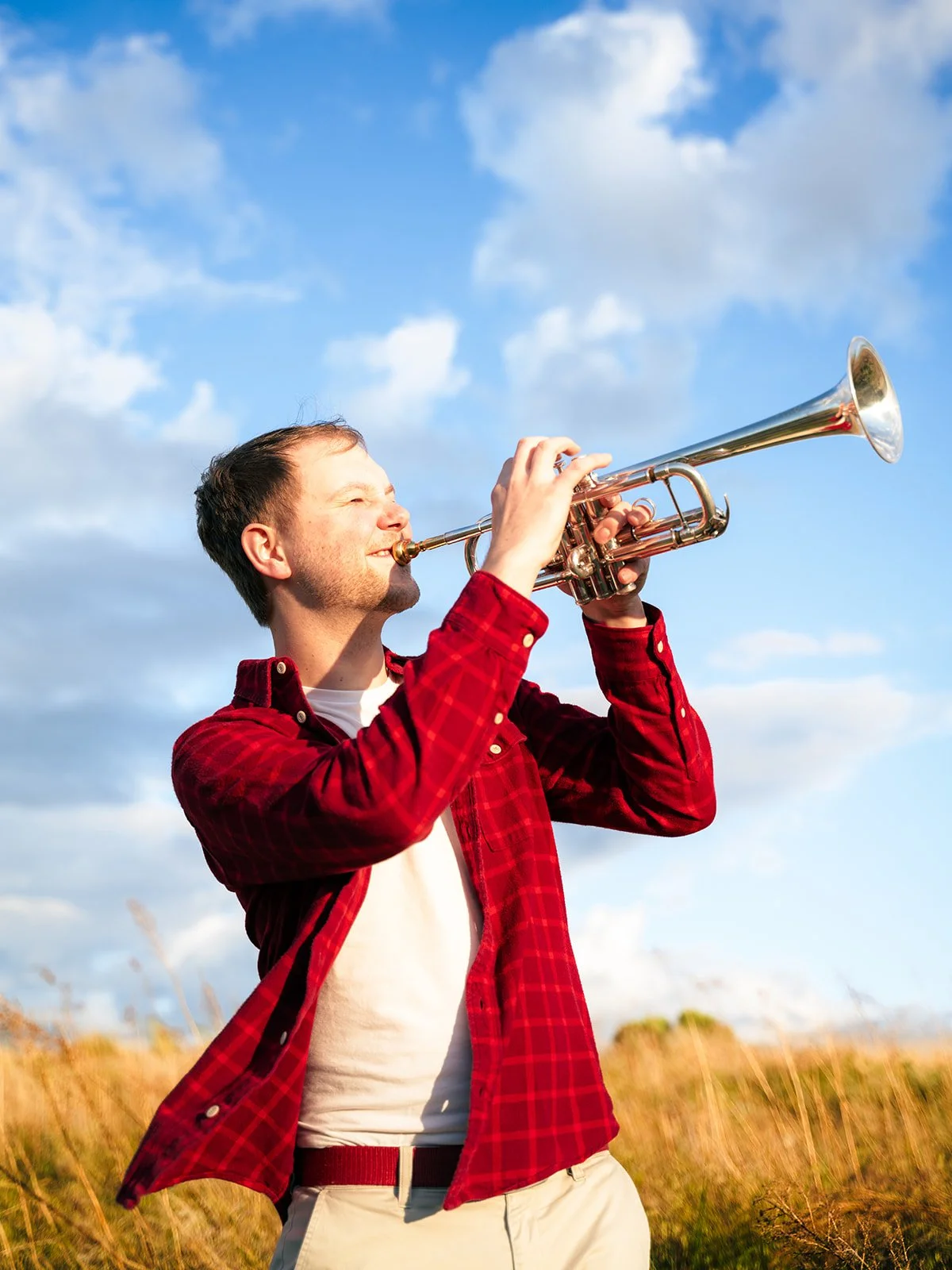 A man in a red plaid shirt playing a trumpet outdoors on a sunny day with blue sky and clouds.