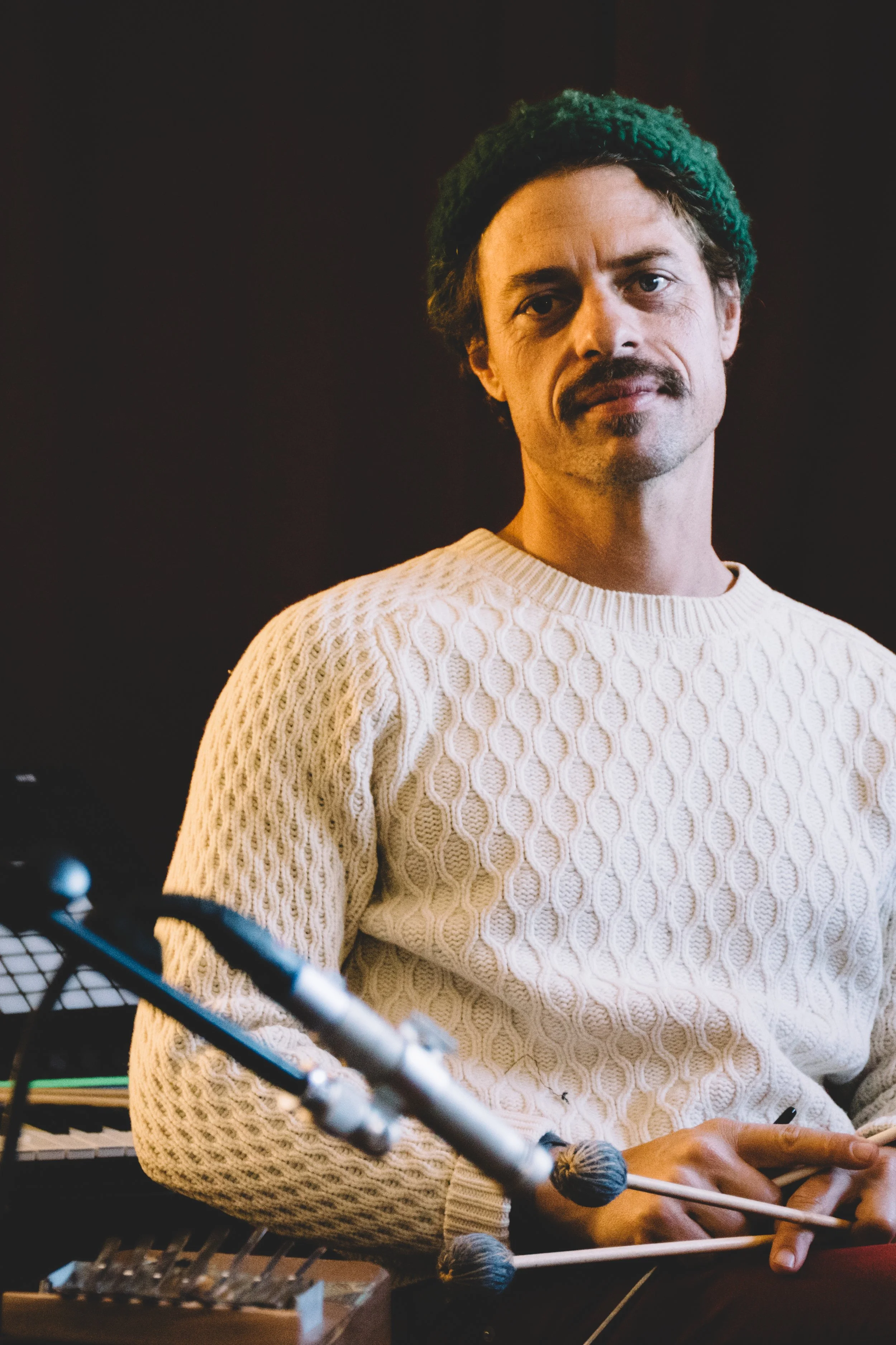 A man with curly hair, a mustache, and goatee wearing a green beanie and a white textured sweater, holding mallets, sitting next to a marimba.