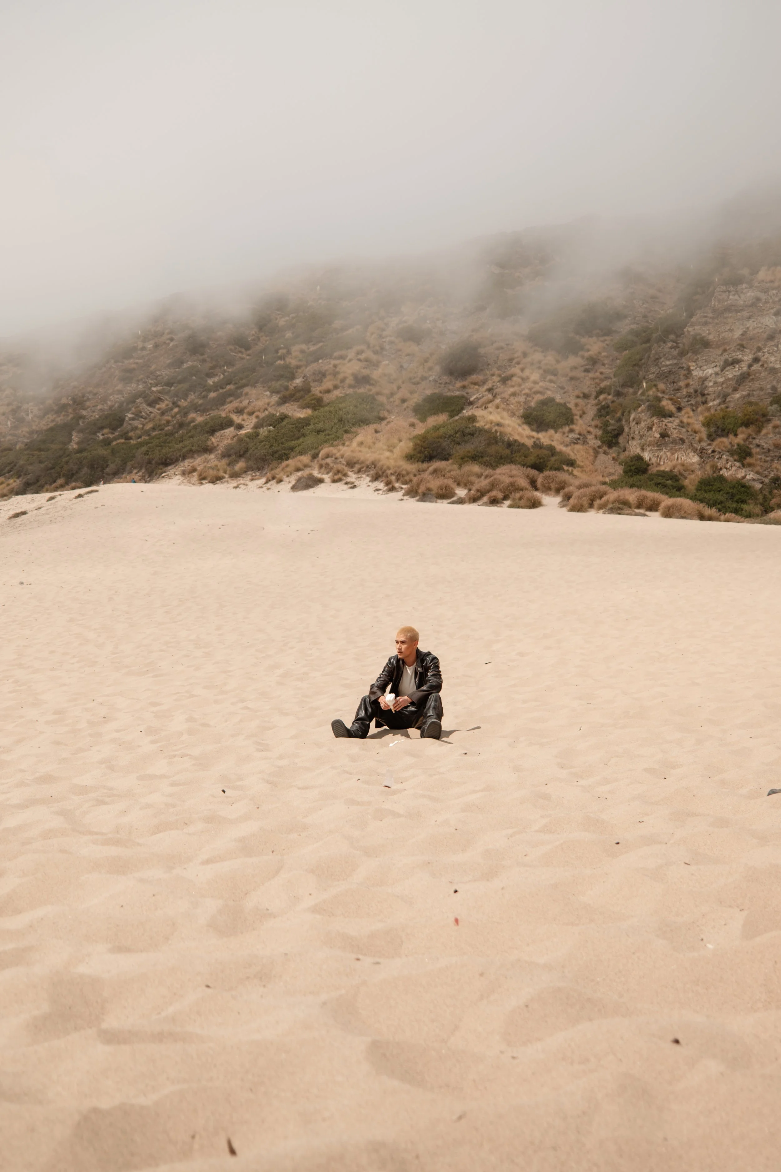 A person sitting alone in the middle of a sandy beach with hills and fog in the background.