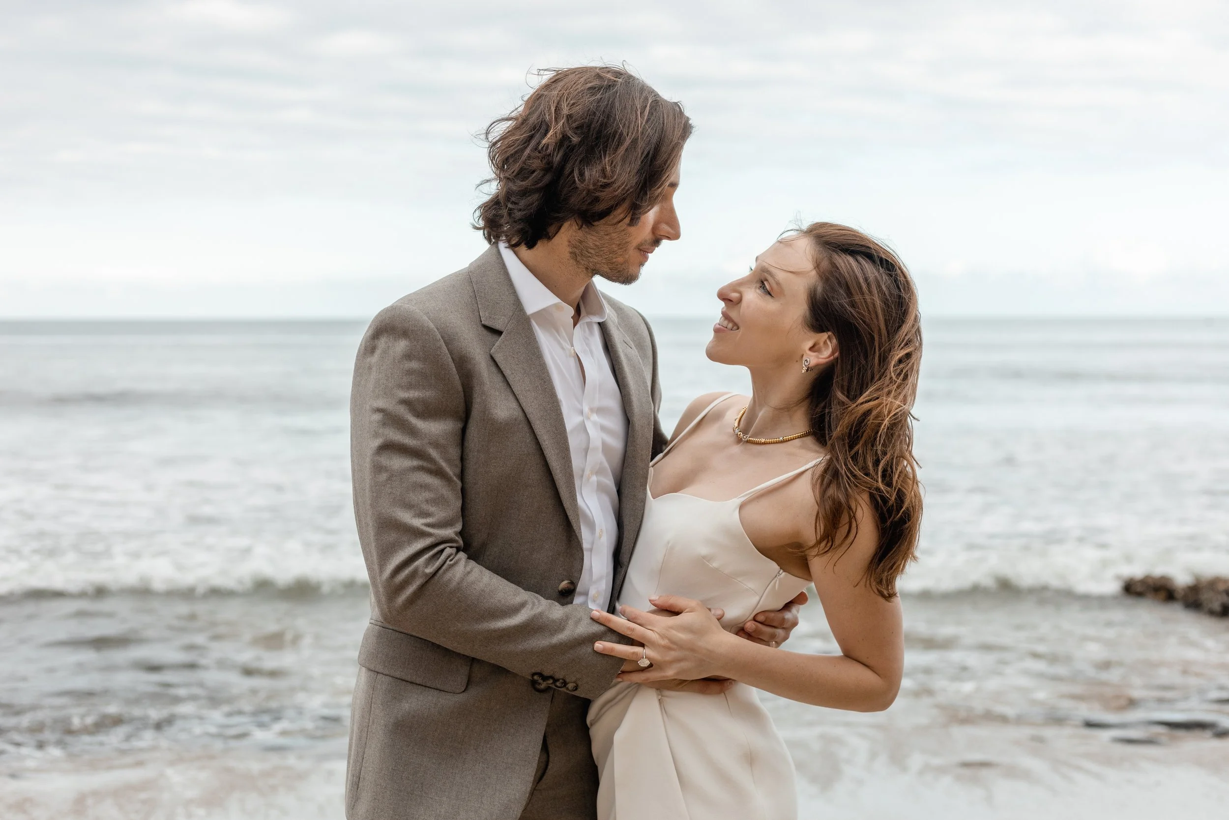 A couple in formal attire stands closely on the beach, gazing into each other's eyes with the sea and sky in the background.