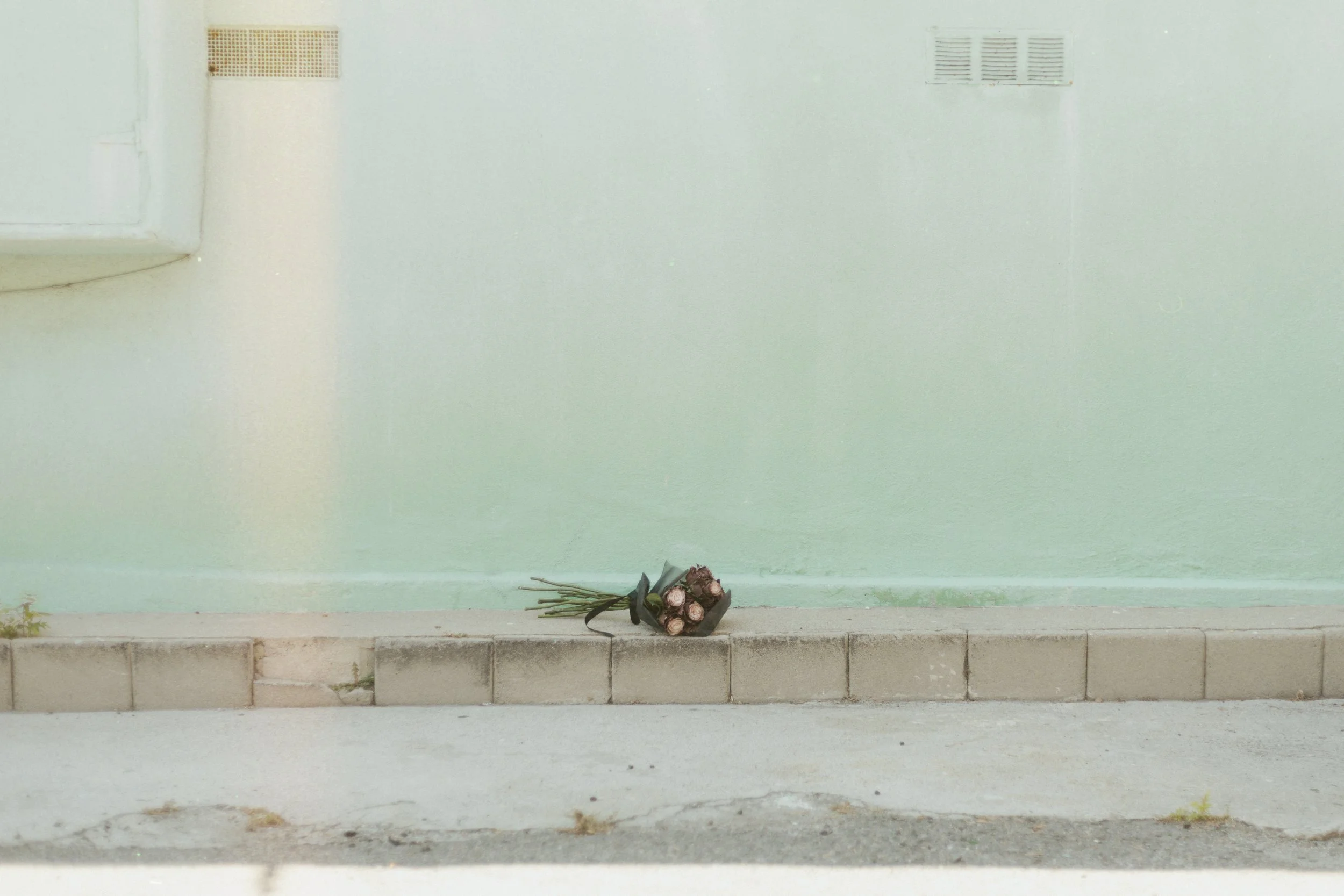 A bouquet of pink roses wrapped in black paper and tied with a black ribbon, resting on a concrete sidewalk against a pastel green wall with vents.