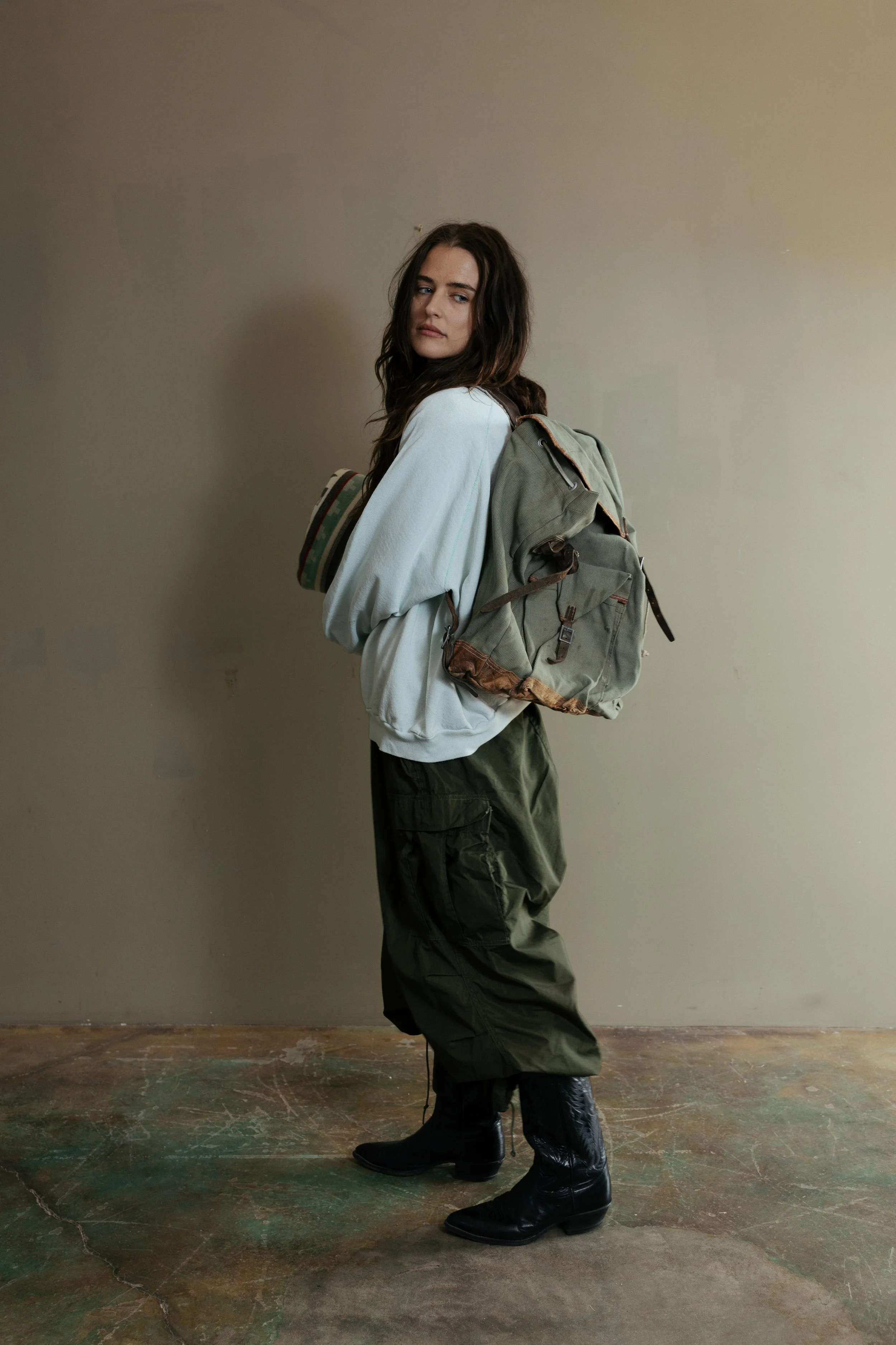 A young woman with long dark hair wearing a white oversized sweatshirt, cargo pants, and black boots, standing against a plain beige wall with a backpack on her back.