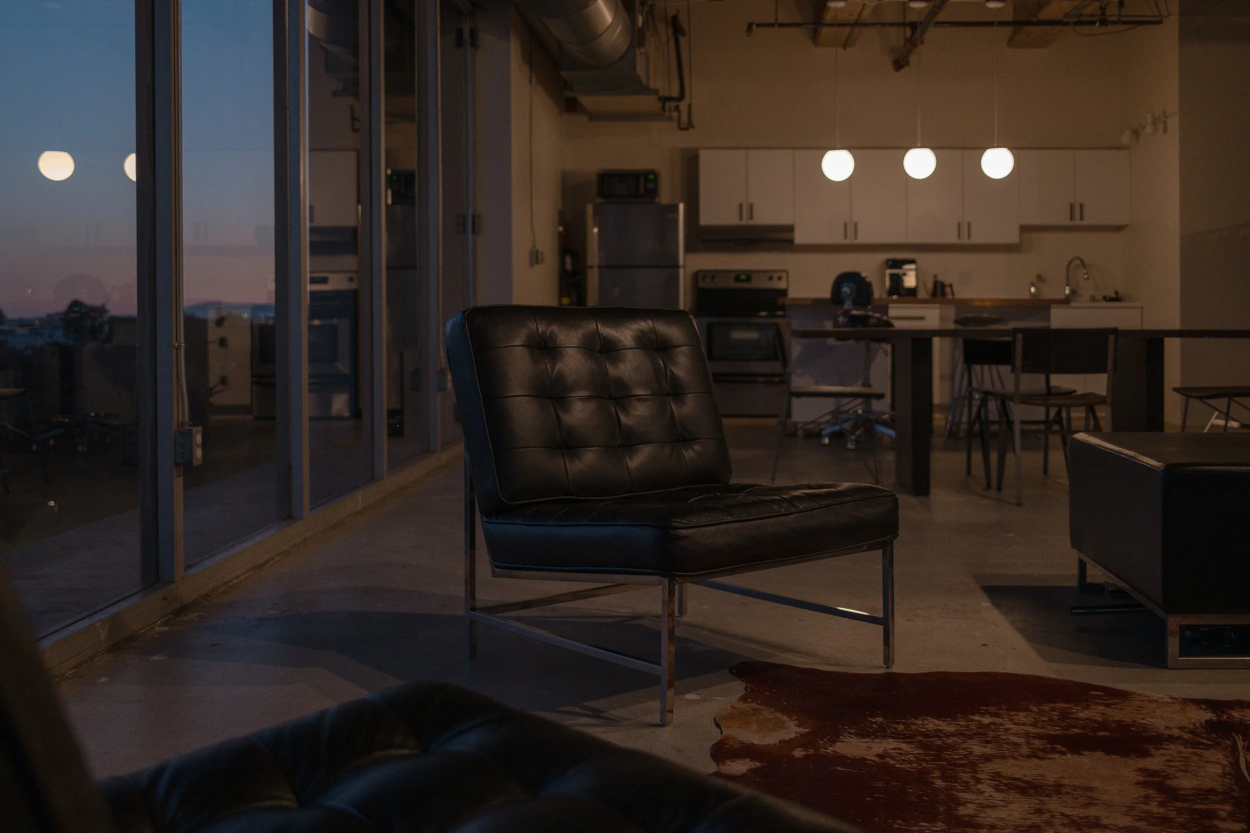 Modern apartment with black leather chair, kitchen in background, and city view through large glass windows.