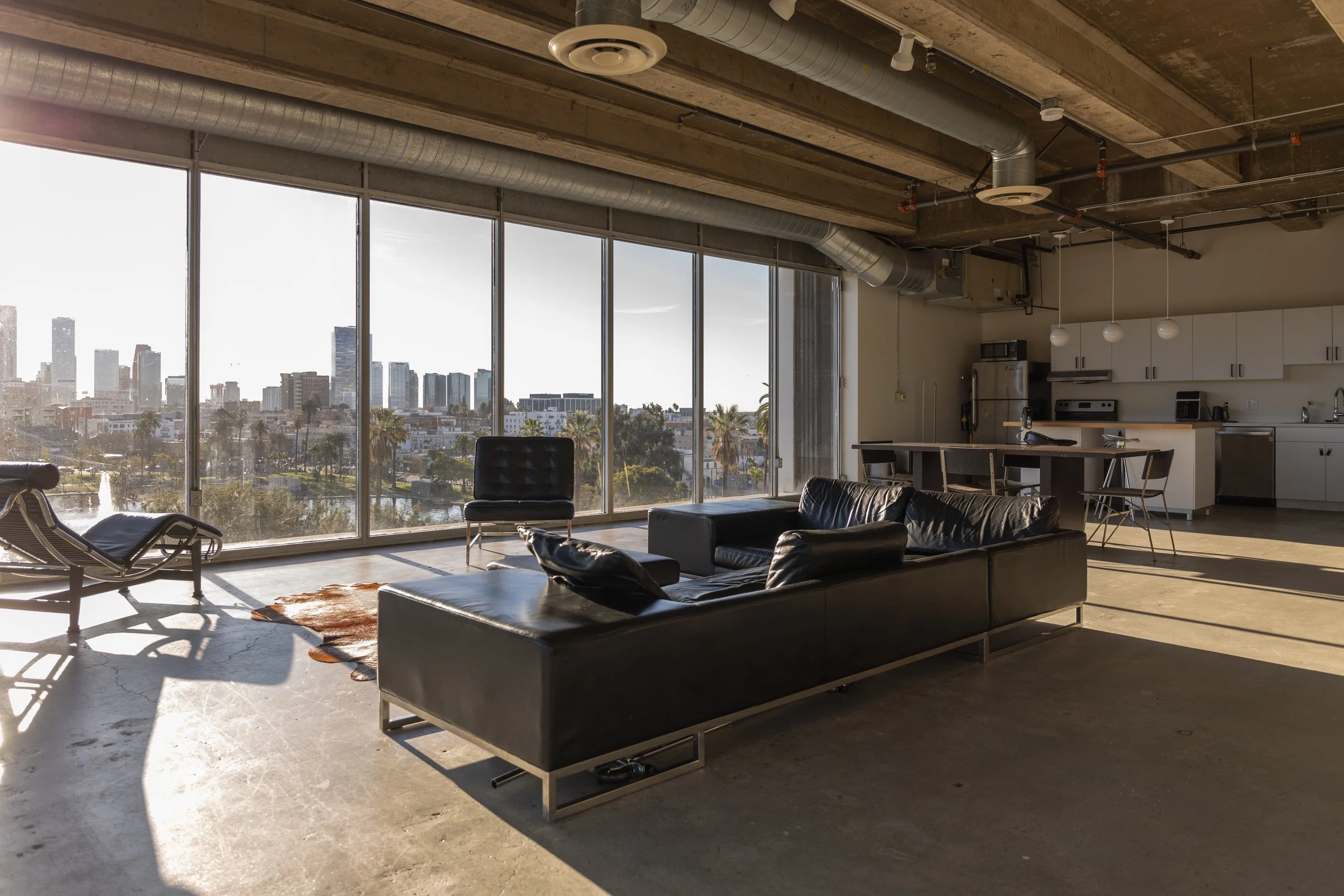 Open-concept living room with large floor-to-ceiling windows overlooking a city skyline, furnished with black leather sofas, an armchair, a coffee table, and a kitchen area with white cabinets, barstools, and pendant lighting.