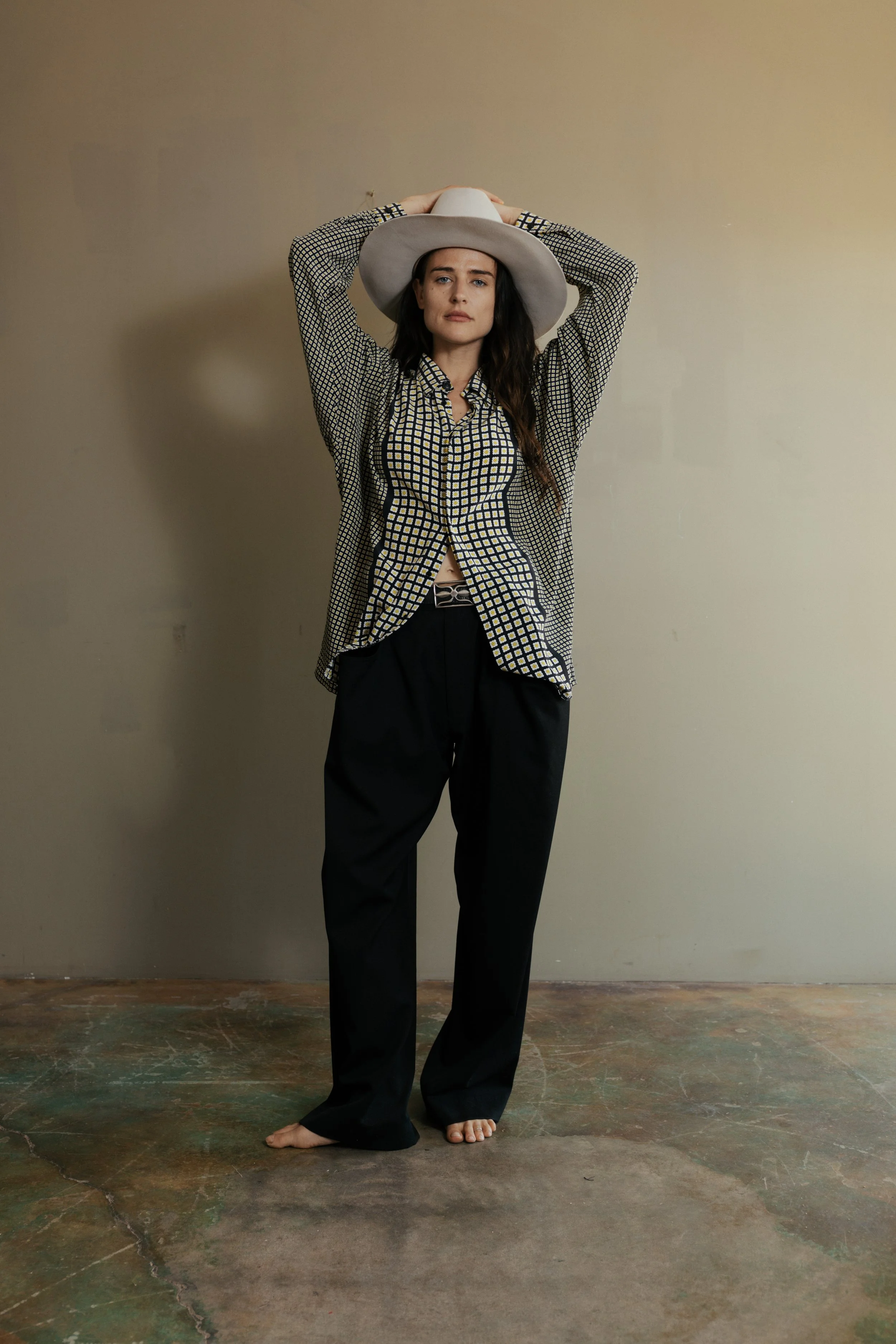 Woman wearing a wide-brimmed hat, patterned blouse, and black pants standing barefoot against a plain wall.