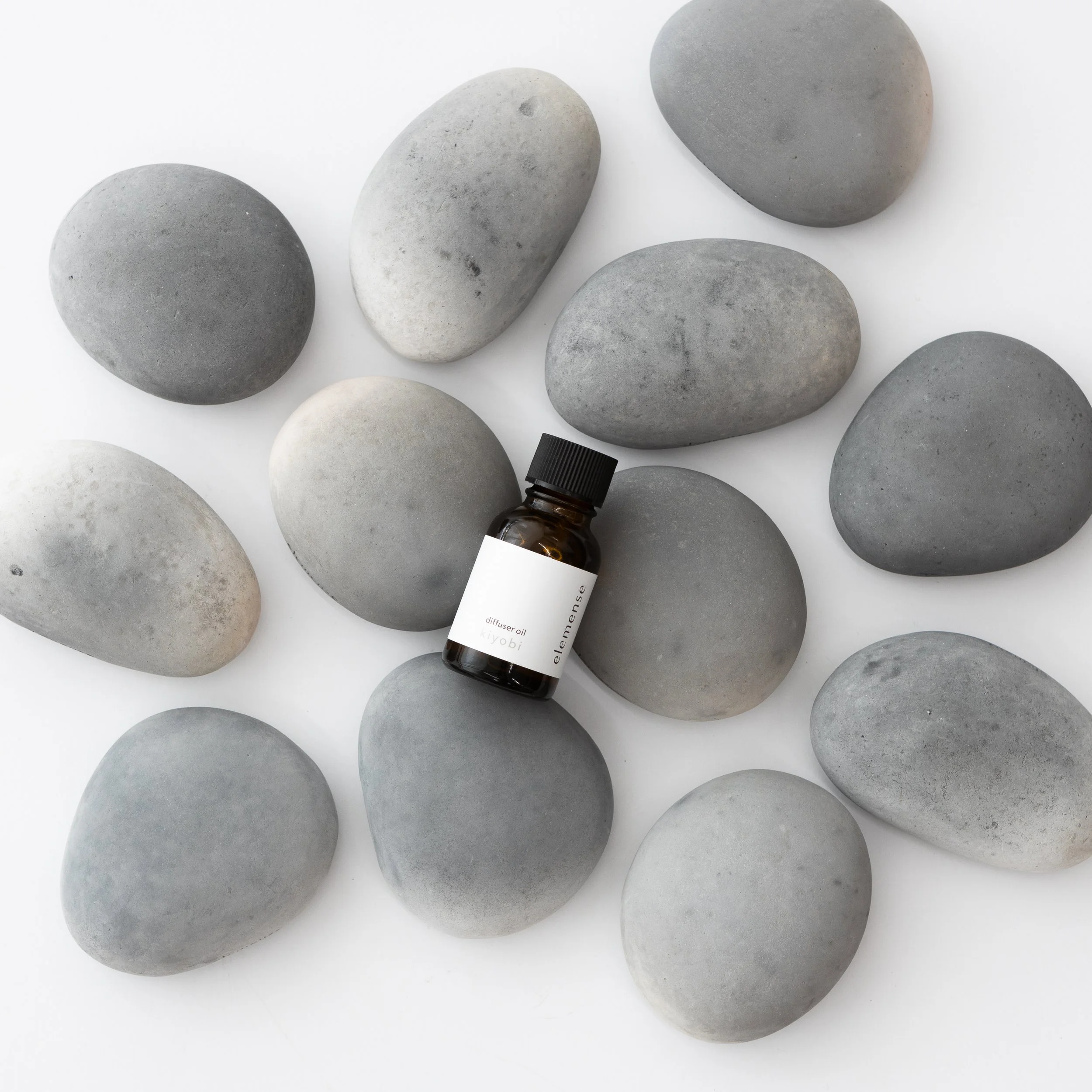 A small brown glass bottle labeled 'diffuser oil kiyobi' resting among smooth, rounded gray stones on a white background.