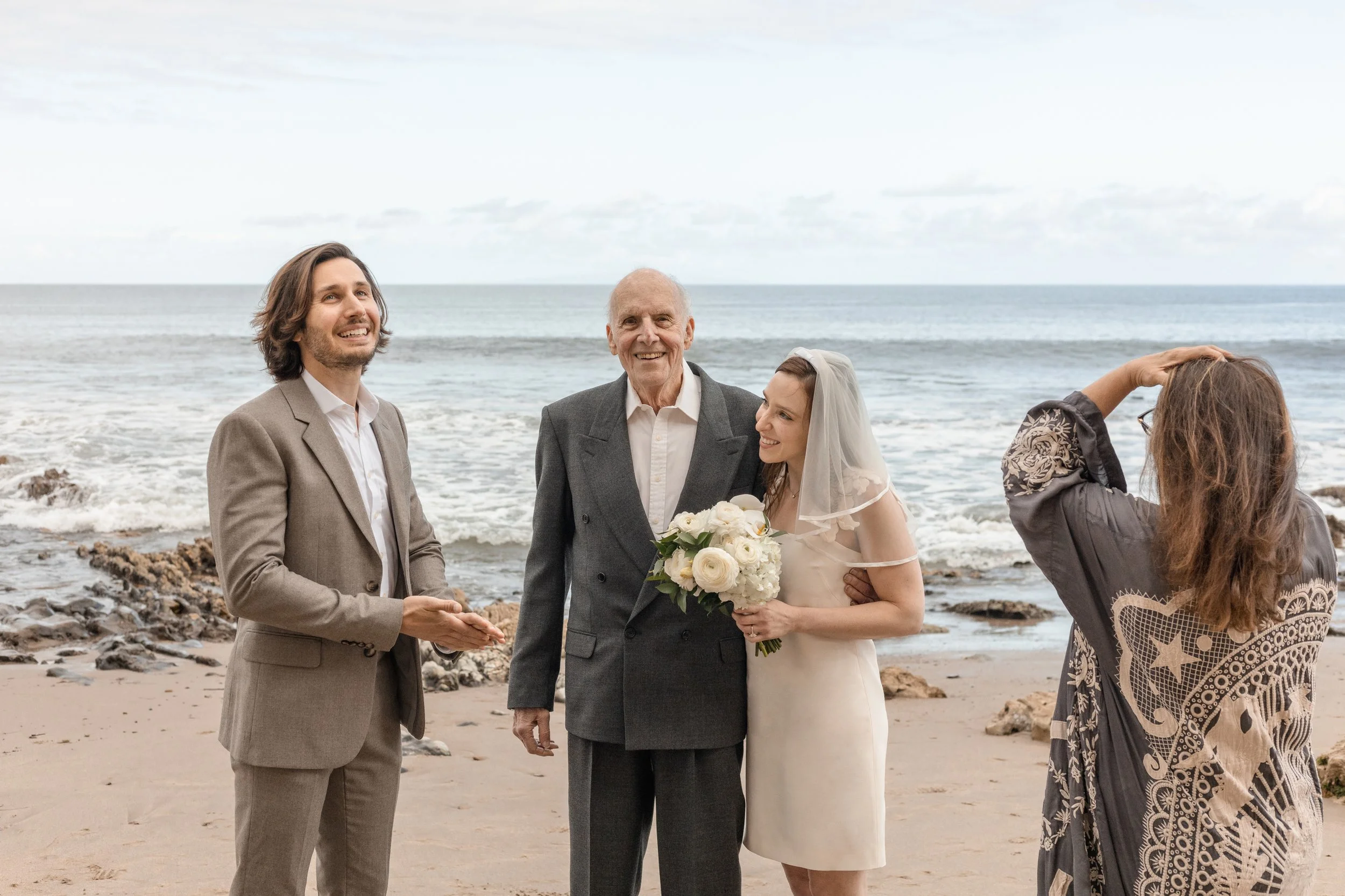 A wedding ceremony taking place on a beach with four people: a bride holding a bouquet of flowers, an older man in a gray suit, a groom in a tan suit, and a woman adjusting her hair. The ocean is in the background.