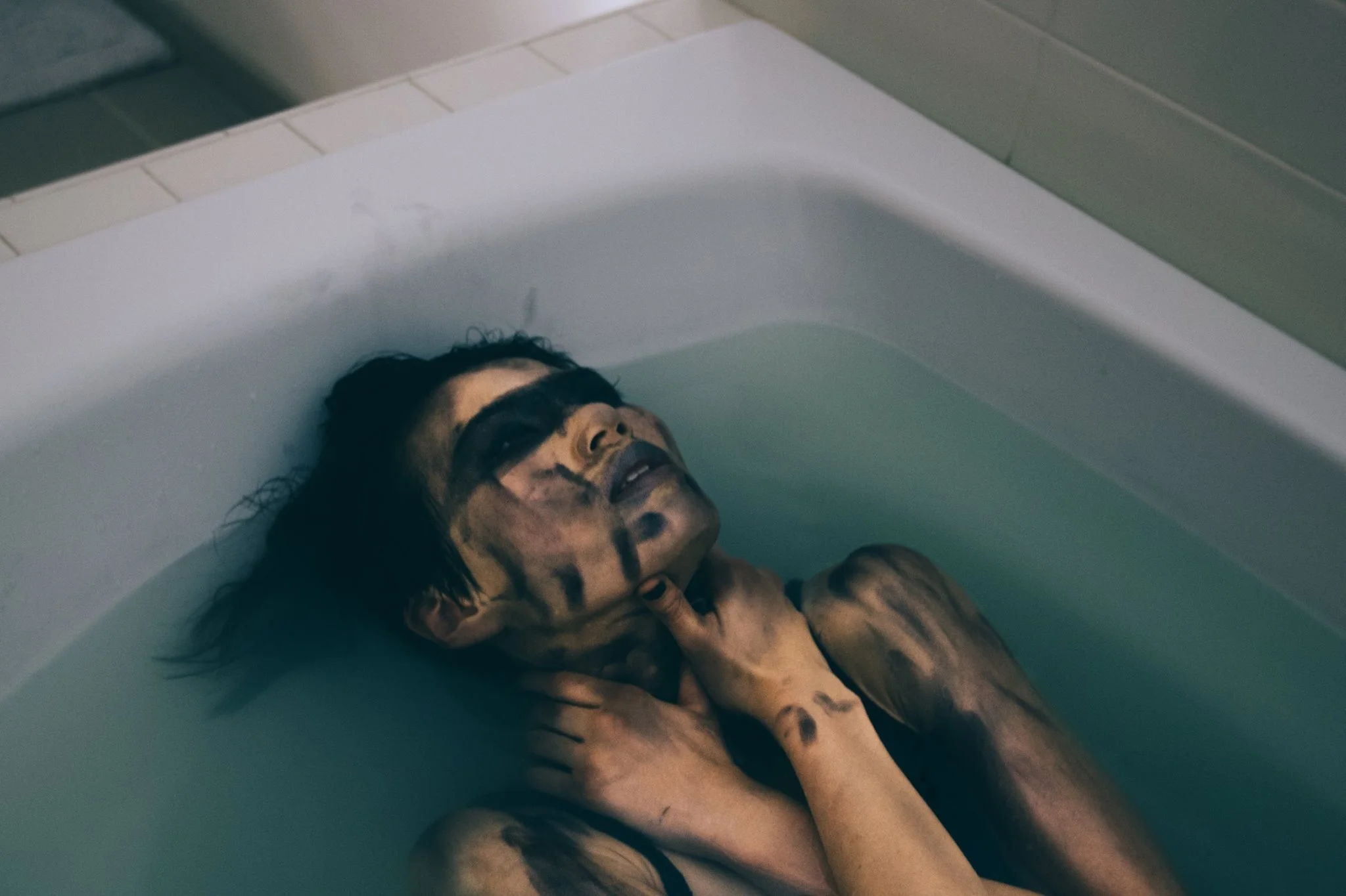A person with dark hair lying in a bathtub filled with water, covered in black and dark colored paint or makeup, with their hands near their neck and face, looking upward.