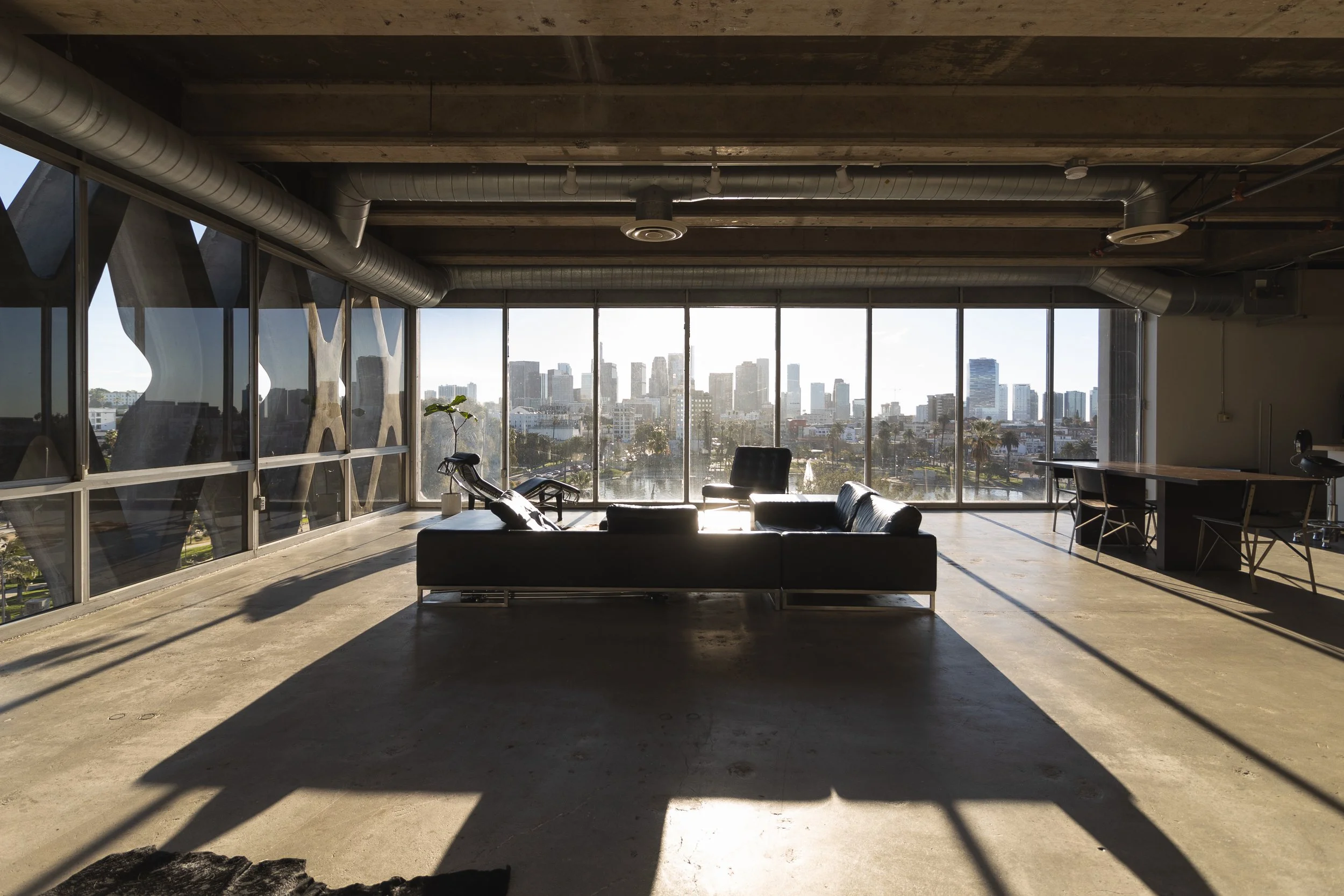 Modern office lounge with large windows showing a city skyline, black leather couch, desk, chair, and indoor plant