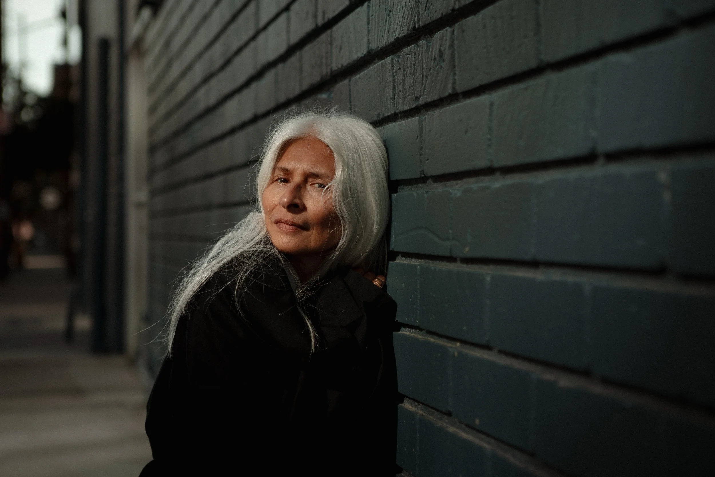 An older woman with long white hair leaning against a dark brick wall on a city street at night.
